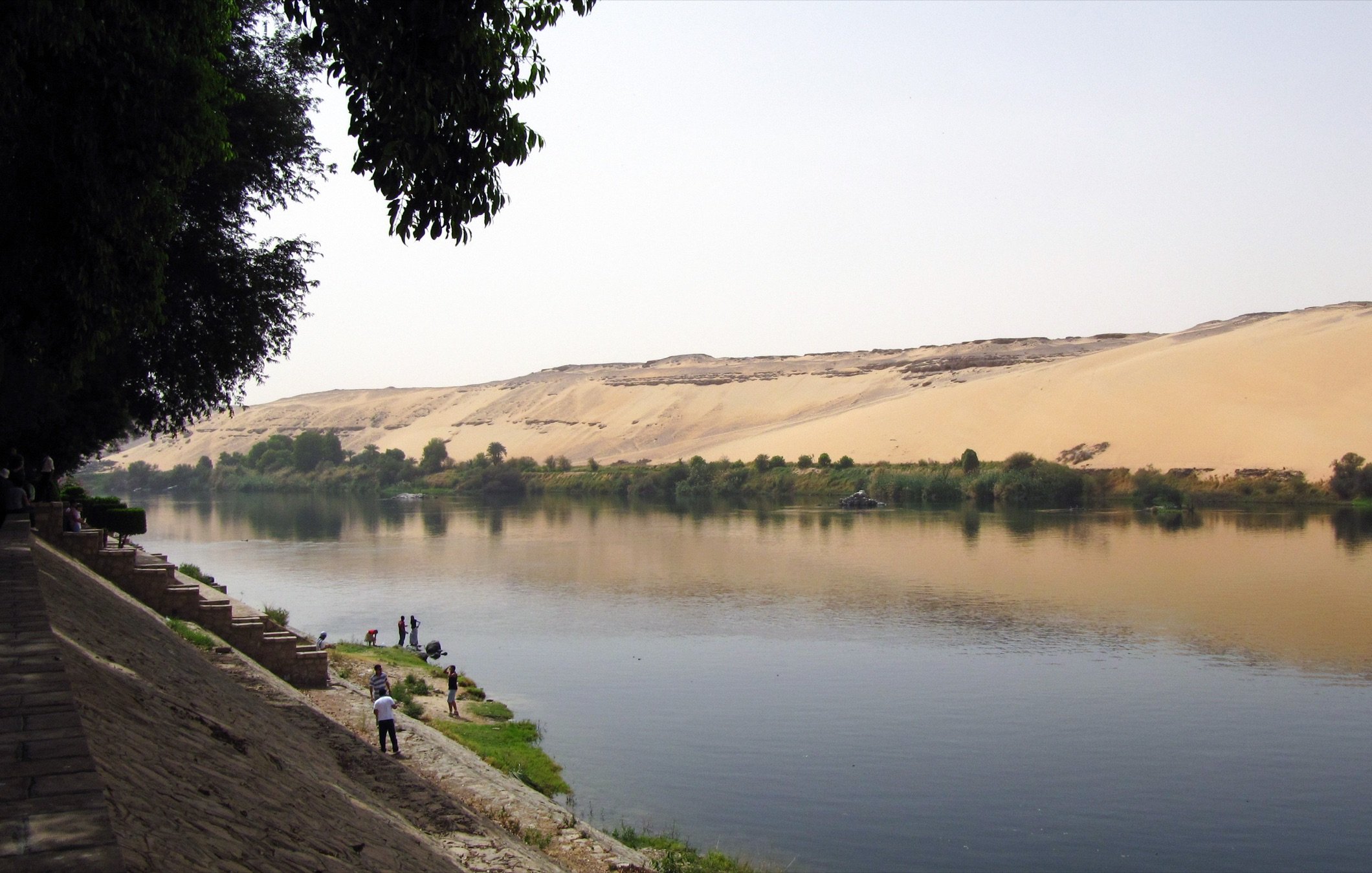 L'image montre un paysage paisible où un fleuve serpente le long d'une rive. Sur un côté, il y a des dunes de sable doré qui montent en pente, créant un contraste avec la verdure des arbres qui bordent le fleuve. Des personnes se trouvent au bord de l'eau, peut-être en train de pêcher ou de profiter de la nature. Le ciel est clair, et la lumière dubaine donne une atmosphère sereine à la scène.