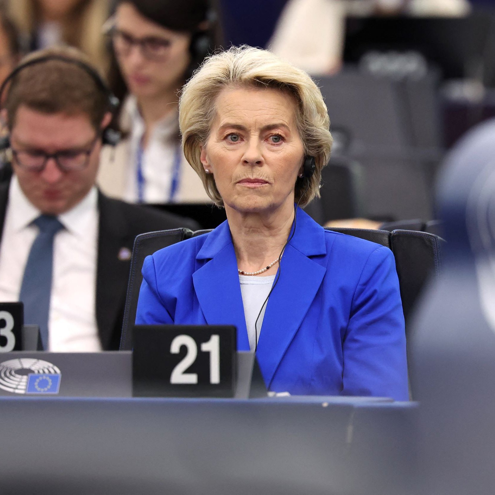 در این تصویر، یک زن با موی بلوند و کوتاه که لباس آبی رنگی به تن دارد، در یک جلسه رسمی نشسته است. او به نظر می‌رسد که به سخنرانی یا ارائه‌ای در حال گوش دادن است. در پس‌زمینه، تعدادی از افراد دیگر نیز مشغول به فعالیت هستند و برخی از آن‌ها هدفون به گوش دارند. فضای کلی تصویر رسمی و جدی است.