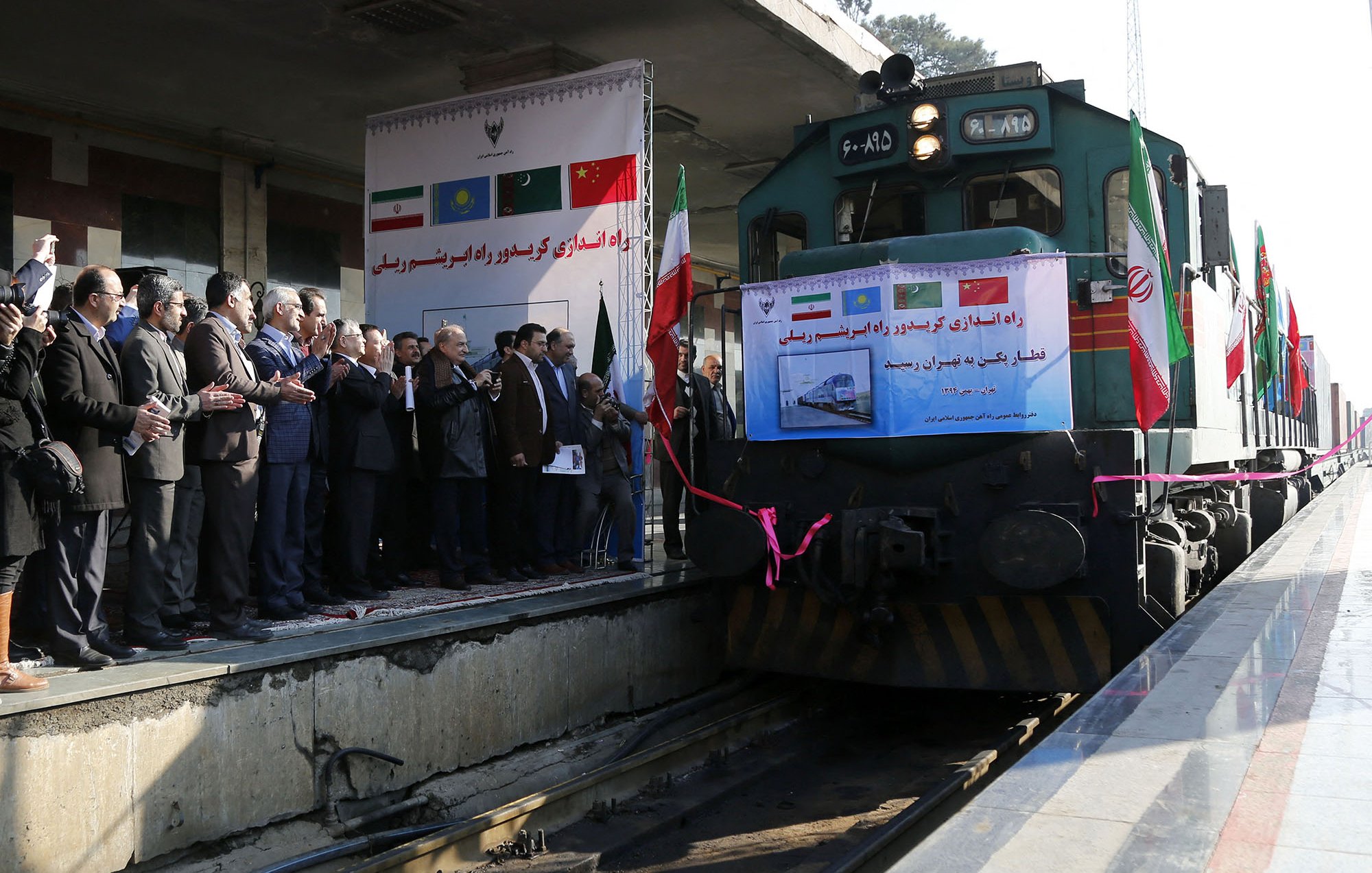 L'image montre un train à quai, avec plusieurs personnes rassemblées sur la plateforme. On peut voir des bannières en arrière-plan, indiquant probablement un événement officiel ou une inauguration. Les participants semblent en train de célébrer quelque chose, avec des applaudissements et des sourires. Le train lui-même est vert et noir, et l'atmosphère semble festive et sombre, suggérant un projet important lié aux transports ou aux liaisons ferroviaires.