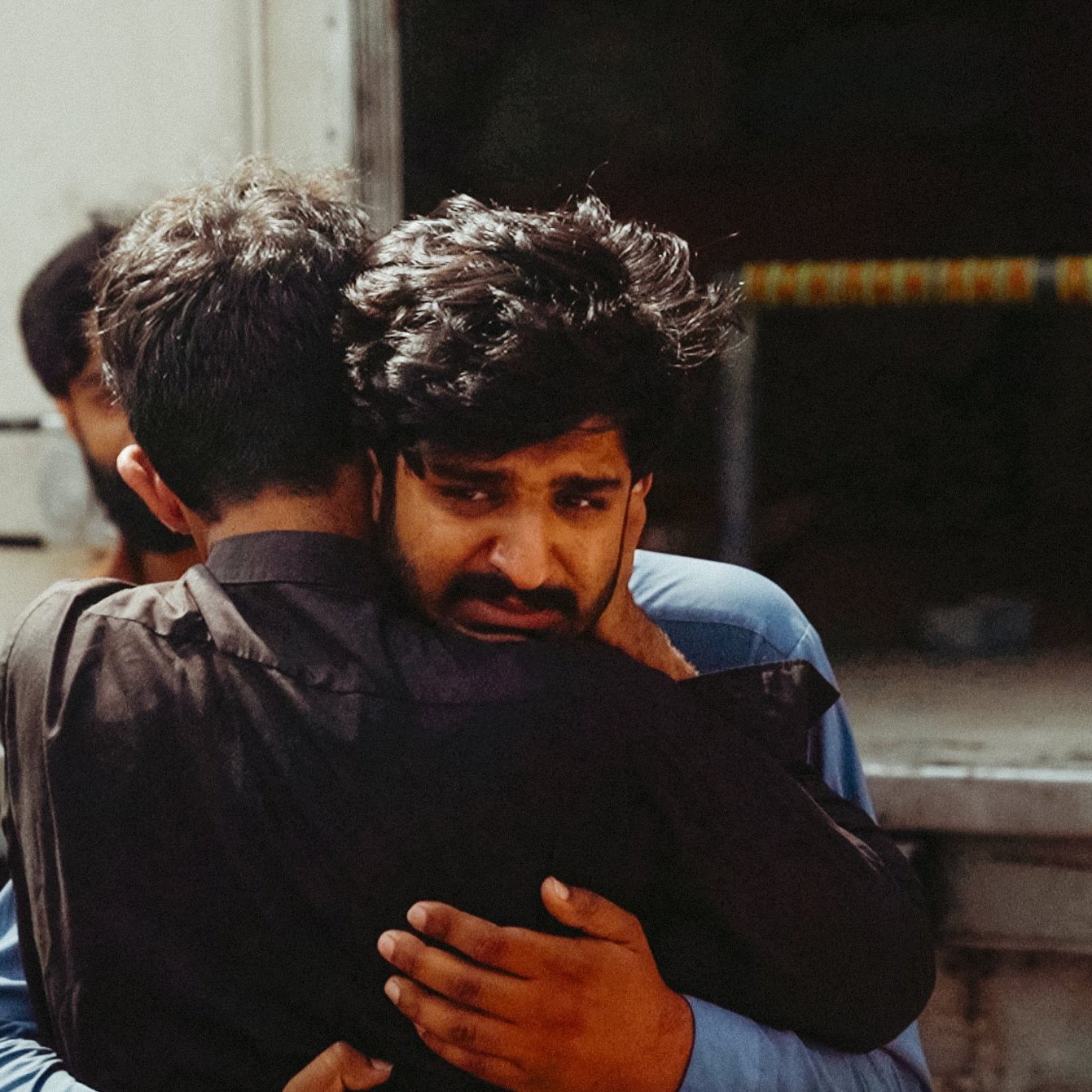 Two men embrace tightly, one looks visibly emotional, surrounded by others in a somber setting.