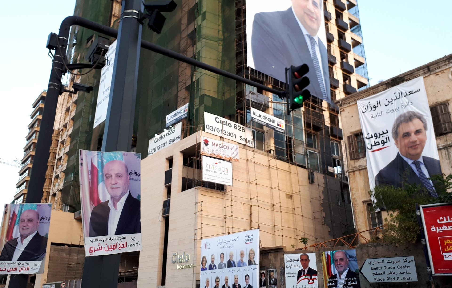 L'image montre un immeuble en construction ou rénové, arborant plusieurs affiches et panneaux publicitaires. Les affiches présentent des portraits de différentes personnes, probablement des candidats ou des figures politiques, accompagnés de textes en arabe. La scène donne l'impression d'une campagne électorale ou d'une promotion politique. Il y a également un feu de signalisation visible à gauche, ce qui indique que cette image est prise dans un environnement urbain.