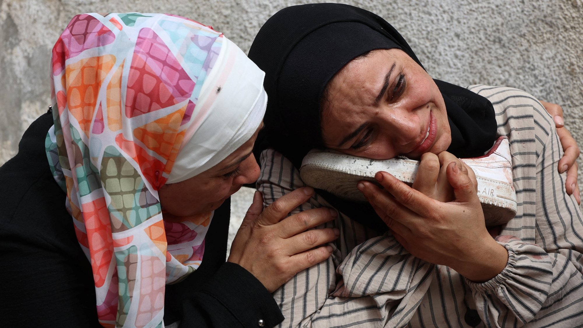 Deux femmes en pleurs, l'une réconfortant l'autre qui tient une chaussure, exprimant une profonde tristesse.