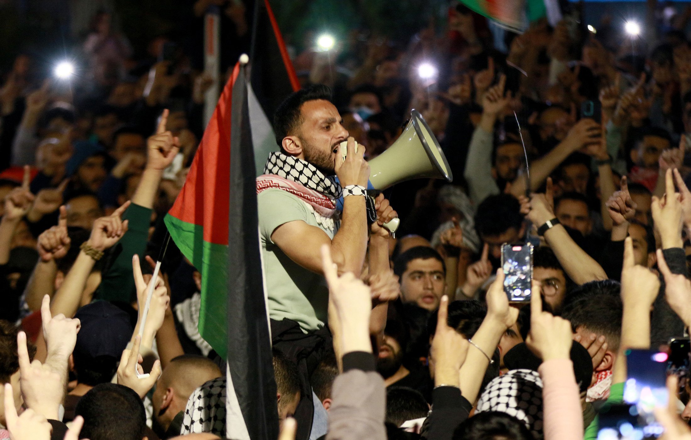 L'image montre un grand rassemblement de personnes, probablement lors d'une manifestation. Au premier plan, un homme utilisant un mégaphone pour parler à la foule. Il est entouré de nombreux participants qui lèvent les bras et brandissent des drapeaux, notamment des drapeaux palestiniens. L'atmosphère semble électrisante et engagée, avec des gens qui filment l'événement avec leurs téléphones. La scène dénote une forte mobilisation et un sentiment collectif.
