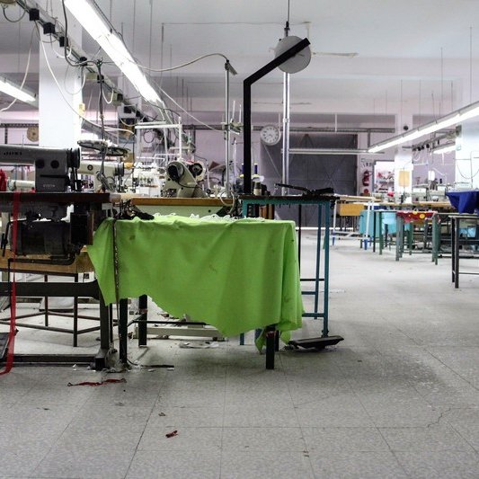 L'image montre un atelier de couture ou de fabrication. On y voit plusieurs tables de travail avec des machines à coudre. Certaines machines semblent être à l'arrêt, et des morceaux de tissu sont éparpillés sur le sol. L'éclairage est assez lumineux, et l'espace semble vaste mais légèrement en désordre, ce qui donne une impression d'activité récente. Des outils et du matériel de couture sont visibles sur les surfaces de travail, et le décor est fonctionnel et utilitaire.
