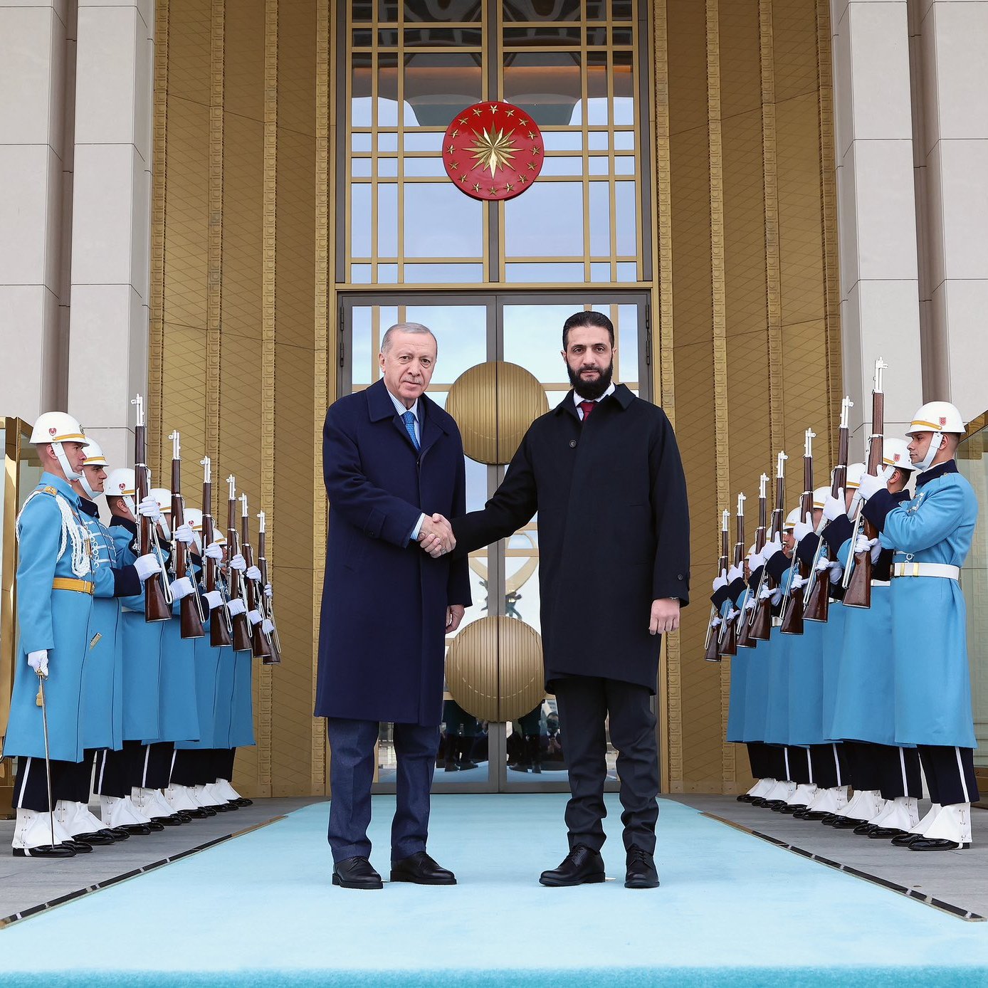 L'image montre deux hommes se serrant la main devant un bâtiment officiel. Ils sont entourés de gardes en uniforme bleu, tenant des fusils, et un grand symbole turc est visible derrière eux. La scène dégage une atmosphère solennelle.