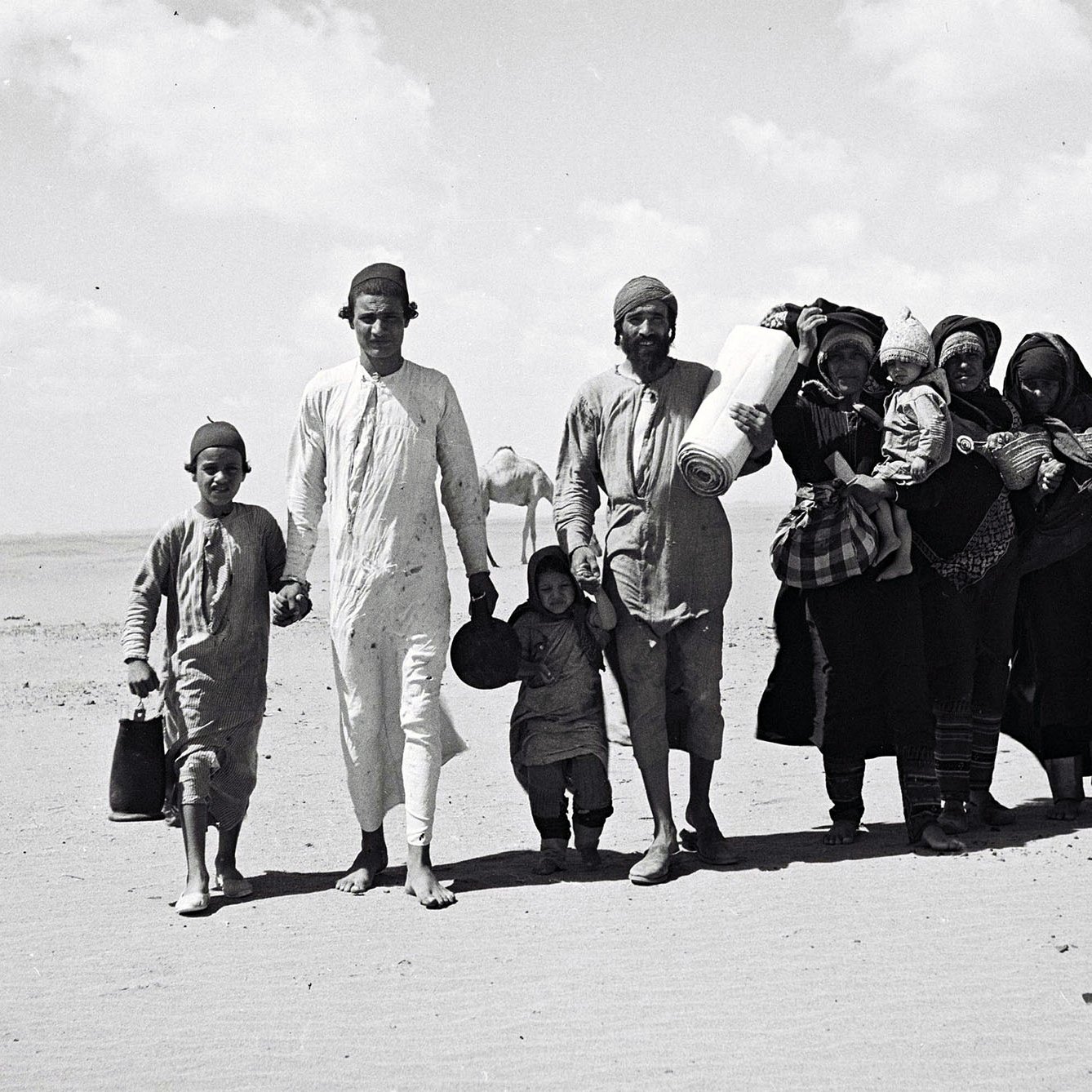 L'image montre un groupe de personnes marchant sur une surface sablonneuse, probablement dans un désert. Au premier plan, on peut voir plusieurs enfants et adultes. Les adultes portent des vêtements traditionnels. Certains membres du groupe transportent des objets sur leurs épaules ou dans les bras, tandis que les enfants semblent tenir la main de l'un des adultes. L'arrière-plan est vaste et dégagé, avec quelques nuages dans le ciel. L'atmosphère dégage une impression de déplacement ou de voyage.