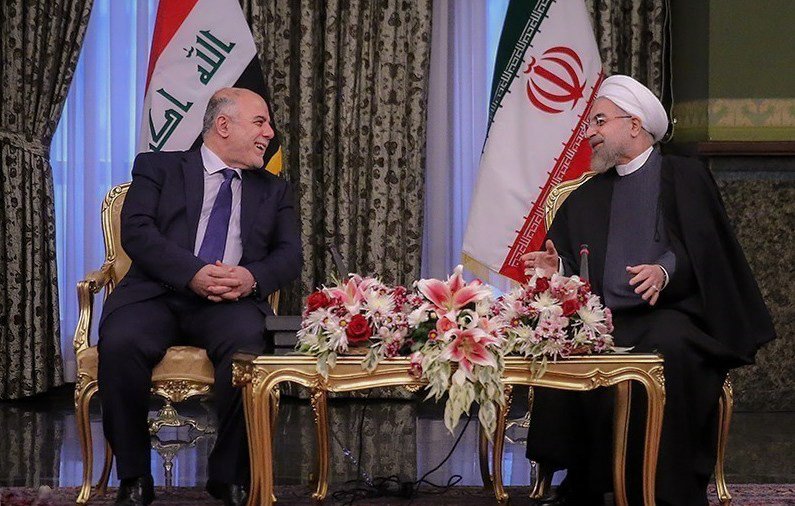 L'image montre une réunion entre deux hommes assis à une table décorée de fleurs. En arrière-plan, on peut voir les drapeaux de l'Irak et de l'Iran. Les deux personnages semblent en discussion amicale, avec des sourires et une ambiance conviviale. Le décor est élégant, avec des rideaux riches et un mobilier doré.