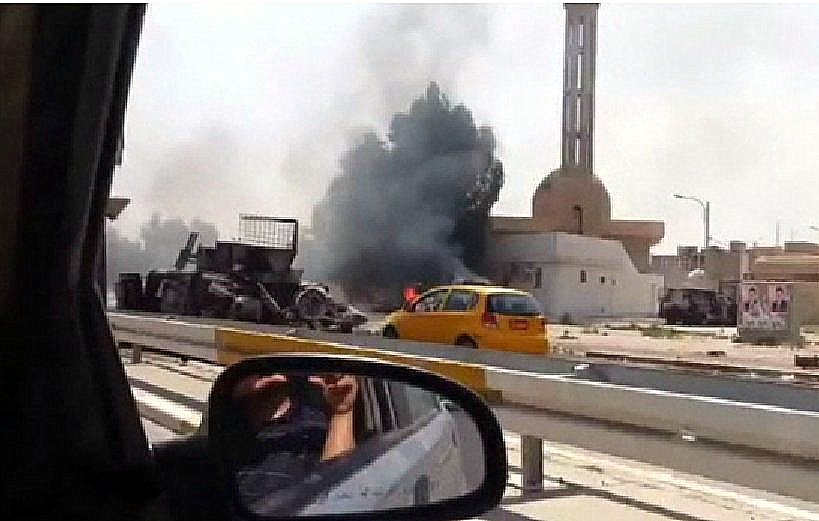 L'image montre une scène d'explosion ou d'incendie, avec de la fumée noire s'échappant d'un bâtiment en arrière-plan. On aperçoit également un véhicule jaune sur la route, tandis qu'une personne semble prendre la photo depuis l'intérieur d'une voiture. Des débris sont visibles près de l'endroit de l'incident, qui pourrait être lié à un conflit. La scène évoque une atmosphère de chaos et de tension.