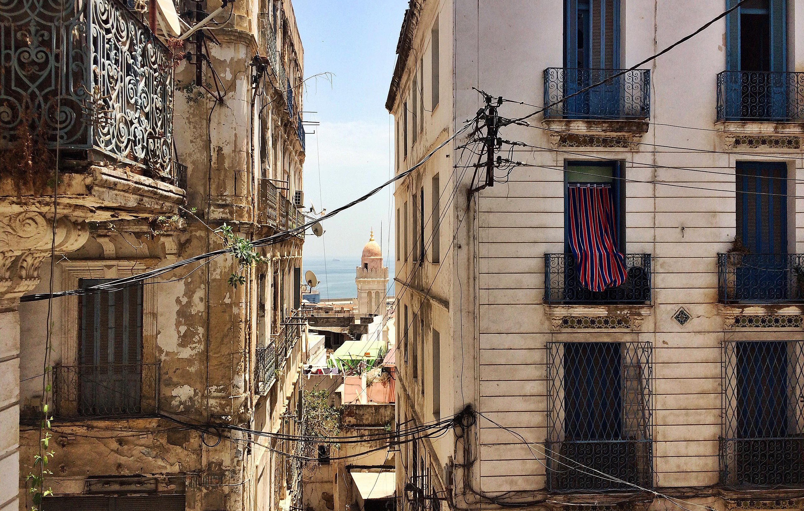 L'image montre une vue d'une rue étroite et animée d'une ville méditerranéenne. De chaque côté, on voit des bâtiments anciens avec des façades usées et des balcons en fer forgé. Des fils électriques serpentent entre les immeubles. Au loin, on peut apercevoir un dôme qui pourrait appartenir à une église ou une mosquée, et un bout de mer est visible, ajoutant une touche de bleu au paysage. Les fenêtres des bâtiments sont ornées de volets ou de rideaux, et l'ensemble dégage une atmosphère nostalgique et pittoresque.