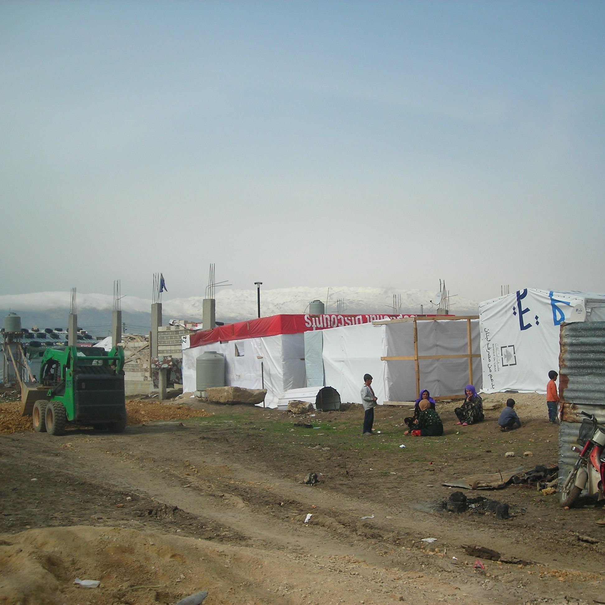 L'image montre un camp de réfugiés ou de personnes déplacées, avec des tentes blanches et des structures temporaires. Il y a un tracteur ou un véhicule de service vert à gauche. Plusieurs personnes sont visibles dans le camp, certaines assises ou debout près des tentes. Le terrain est poussiéreux, et en arrière-plan, on peut apercevoir des montagnes ou des collines. L'ensemble du paysage semble austère et reflète les conditions de vie des occupants. L'atmosphère paraît tranquille, bien que l'environnement soit rudimentaire.
