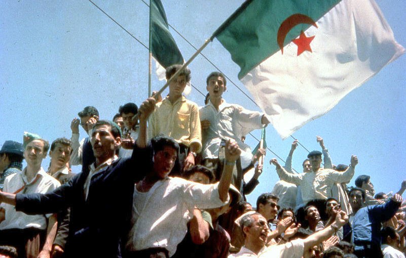 L'image montre une foule rassemblée, levant des bras et brandissant des drapeaux, dont celui de l'Algérie. Les personnes semblent joyeuses et engagées, célébrant un moment fort. L'atmosphère pourrait évoquer une célébration de l'indépendance ou un événement historique significatif pour le pays. Les visages sont animés et l'énergie collective est palpable.