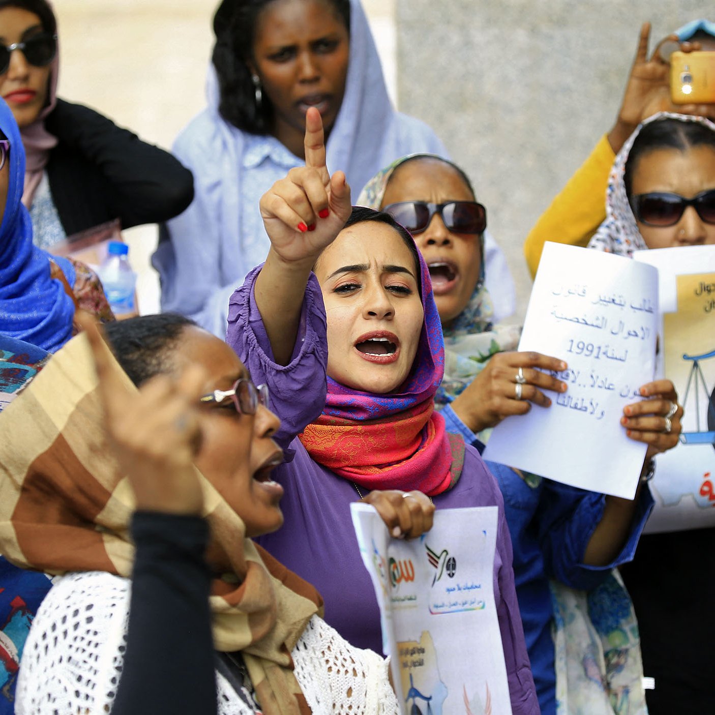 L'image montre un groupe de femmes lors d'une manifestation. Elles portent des foulards et des vêtements colorés. Certaines personnes lèvent les mains et expriment des slogans avec une grande énergie. Des pancartes sont visibles, portant des messages concernant des droits ou des revendications sociales. L'ambiance est dynamique et engagée, avec un fort esprit de solidarité parmi les participantes.