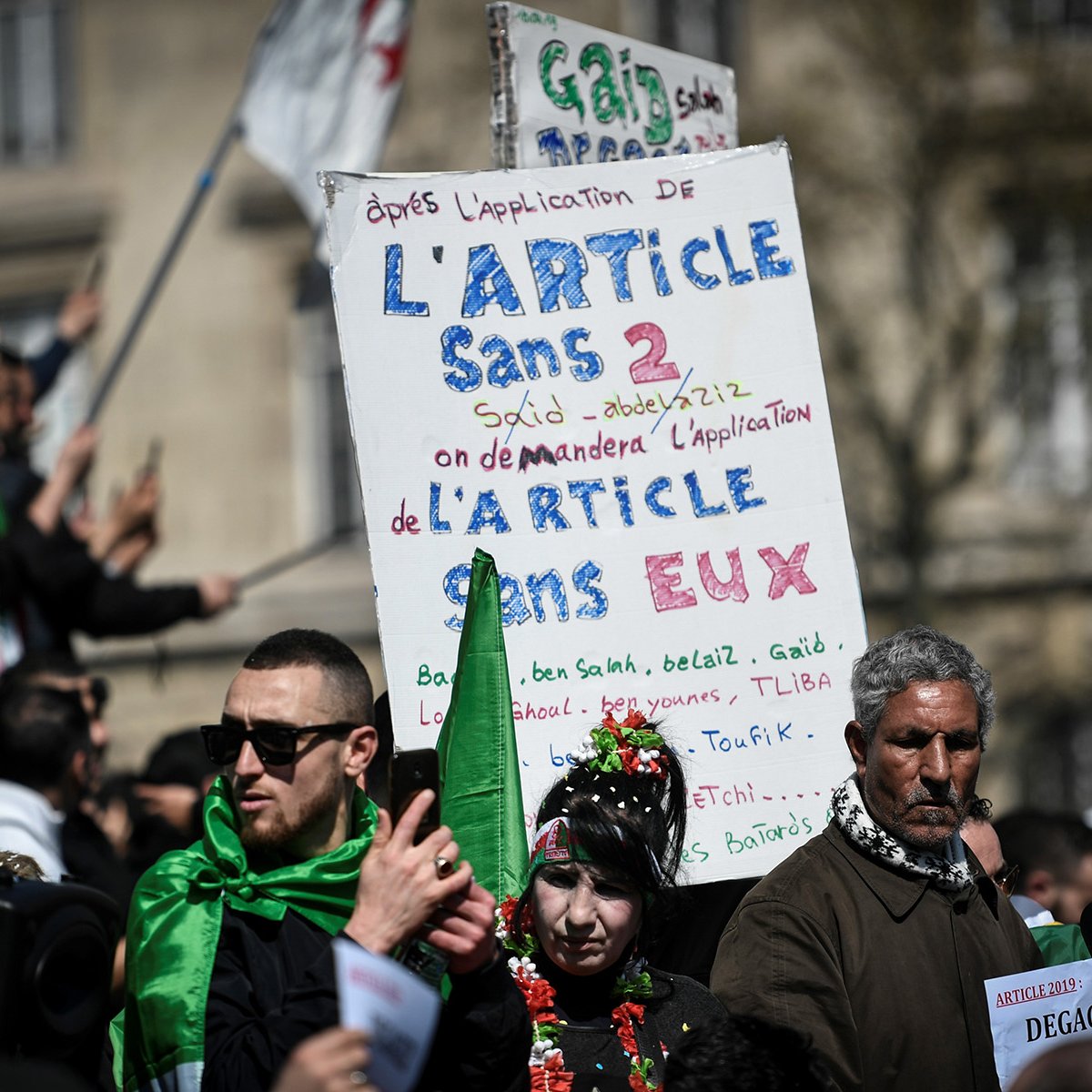 L'image montre une manifestation avec des participants tenant des pancartes. L'une des pancartes est bien visible et contient du texte en français, notamment une référence à l'article 2 et des demandes concernant cet article. Les manifestants portent des vêtements aux couleurs de l'Algérie, ce qui suggère un lien avec des préoccupations politiques ou sociales. L'atmosphère semble sereine mais déterminée, avec des gens rassemblés pour exprimer leurs revendications.