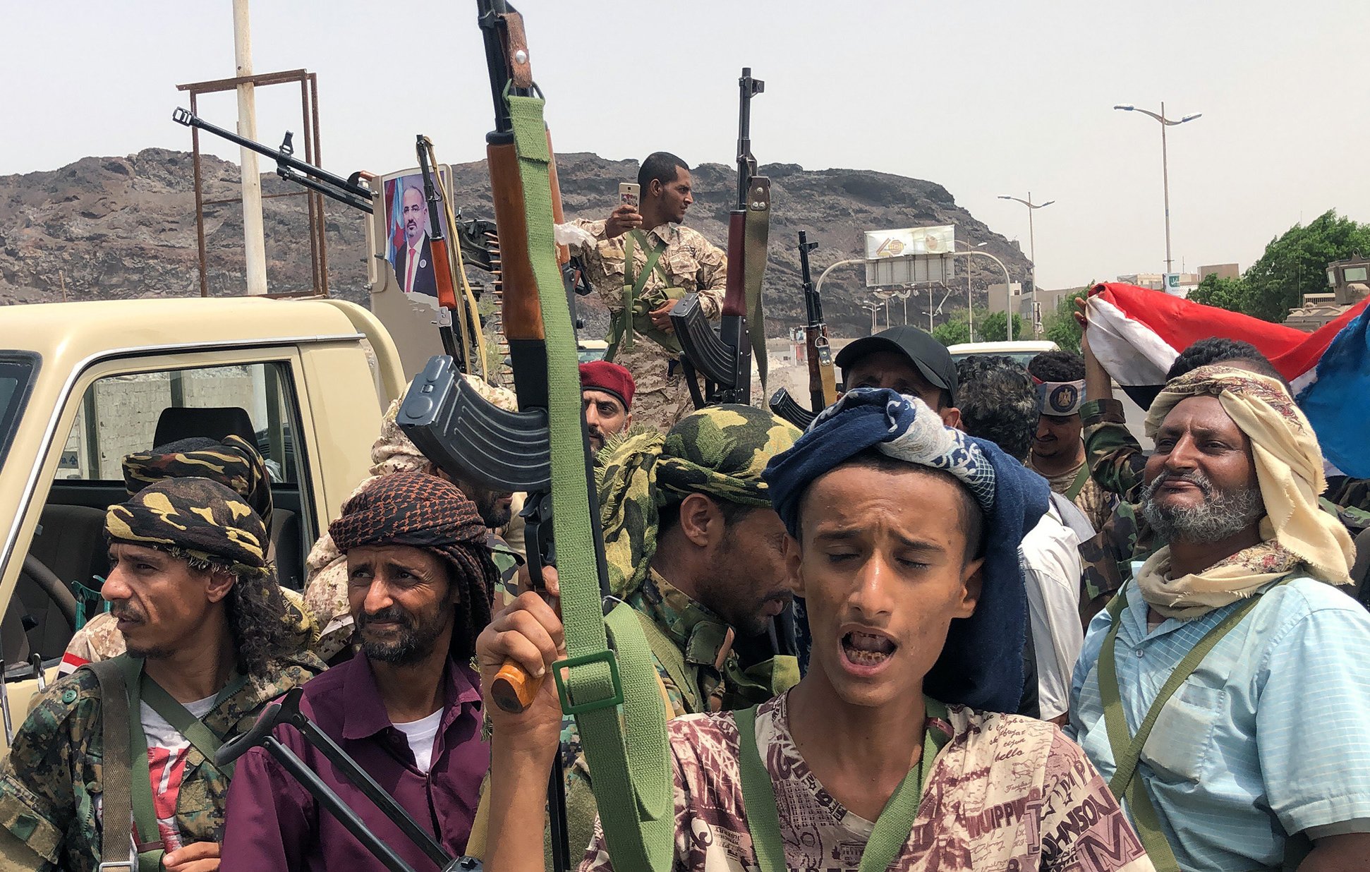 L'image représente un groupe de personnes armées, apparemment réunies dans un contexte de lutte ou de manifestation. Ils portent des vêtements typiques et semblent déterminés, avec des regards engagés. En arrière-plan, on peut apercevoir des montagnes et une circulation modérée, ainsi qu'un drapeau. Un véhicule est également visible, avec des armes visibles entre les membres du groupe. L'atmosphère suggère une situation de tension ou de conflit.