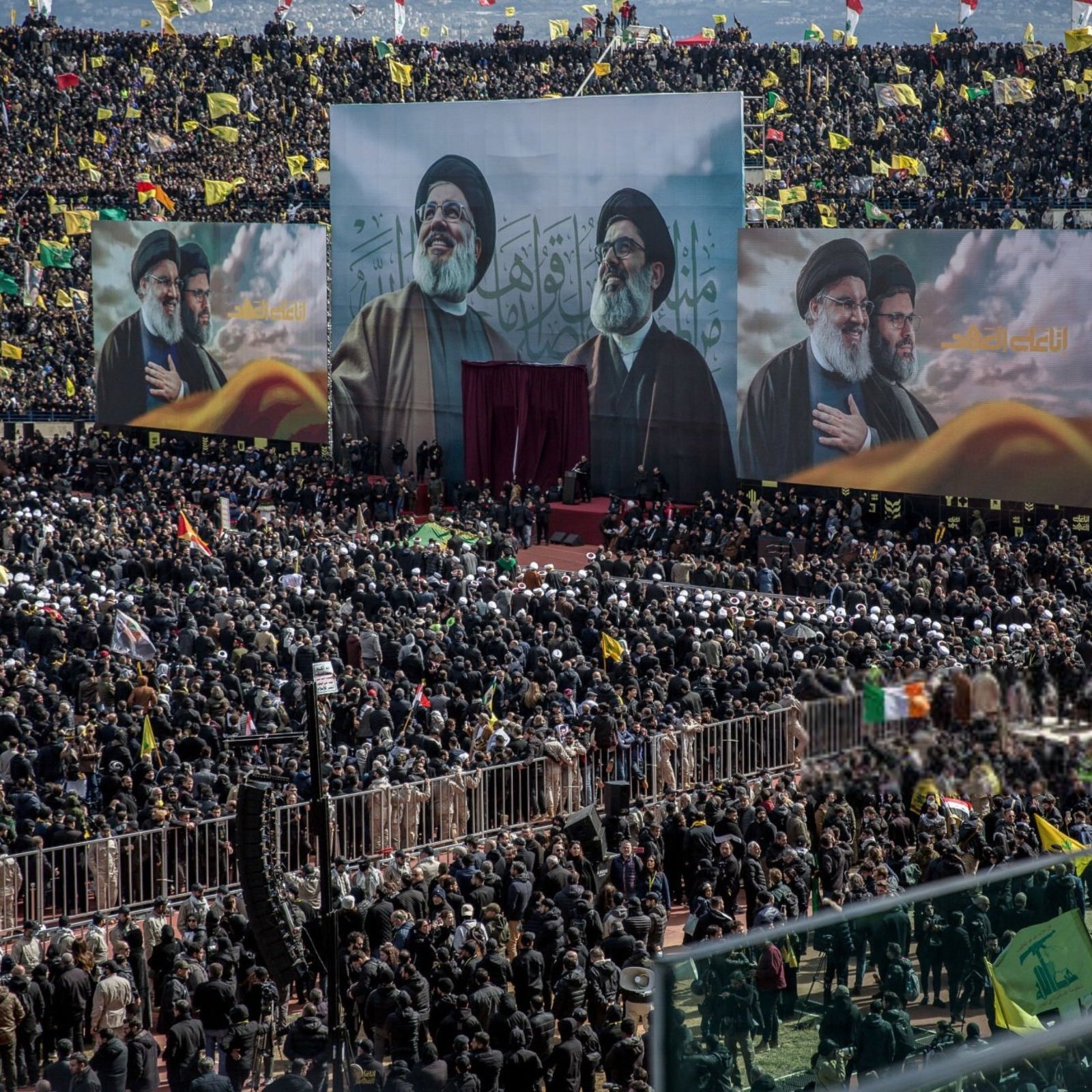 L'image montre une grande foule rassemblée dans un stade, avec des étendues de drapeaux de différentes couleurs. Au centre, trois grandes affiches de figures importantes sont visibles, probablement des leaders religieux ou politiques. L'atmosphère semble festive et engagée, avec des personnes portant des chapeaux ou des foulards spécifiques, suggérant une démonstration de soutien ou de solidarité. Des bannières et des drapeaux supplémentaires peuvent également être aperçus, accentuant l'aspect coloré et vibrant de l'événement.