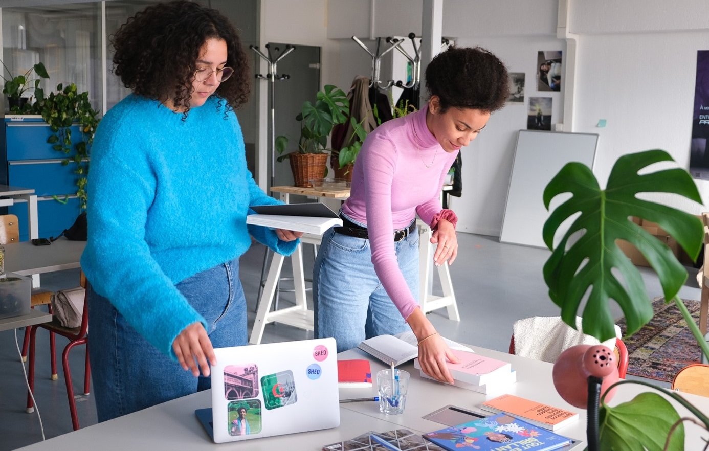 En la imagen se pueden ver a dos mujeres en un espacio de trabajo moderno y luminoso. Una de ellas, con cabello rizado y vestido azul, está sosteniendo unos cuadernos o carpetas, mientras que la otra, con cabello rizado y recogido, está organizando unos libros sobre una mesa. En la mesa hay una computadora portátil, algunos materiales impresos y una planta decorativa. El ambiente parece creativo y acogedor, con elementos de diseño contemporáneo y luminosidad natural.