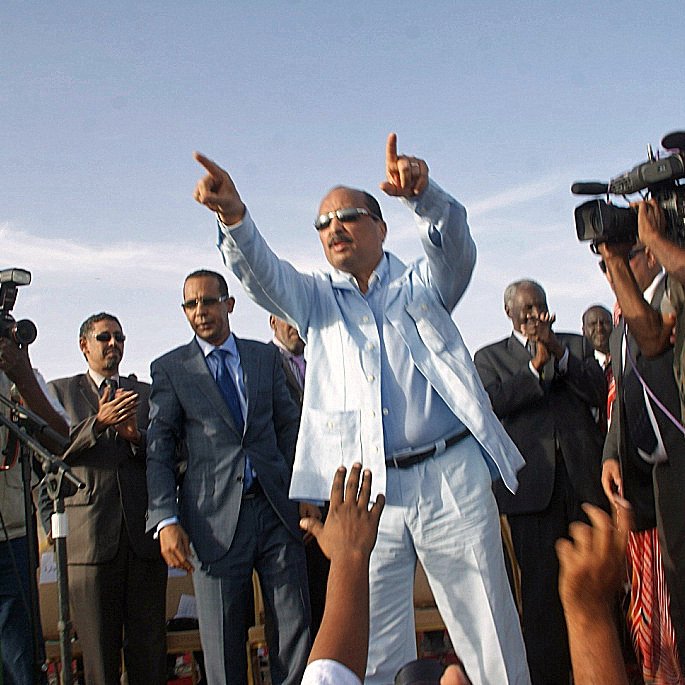 L'image montre un homme debout sur une scène, habillé en costume clair. Il semble être en train de faire un discours et de pointer vers le public. Autour de lui, plusieurs personnes l'écoutent attentivement, dont certaines sont en train de prendre des photos ou de filmer avec des caméras. Il y a une ambiance d'événement public ou politique, avec un discours énergique et engageant.