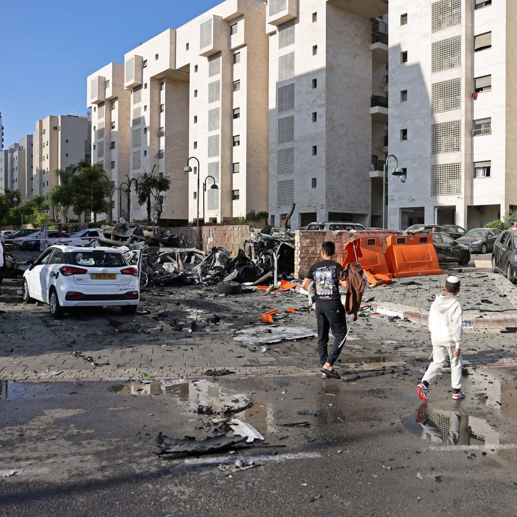 L'image montre une scène de destruction urbaine. On peut voir plusieurs véhicules endommagés, certains complètement détruits. Le sol est couvert de débris, et il semble qu'une explosion ou un événement violent ait eu lieu. En arrière-plan, il y a des bâtiments modernes. Des personnes marchent sur la route, apparemment en train d'évaluer les dégâts. Le ciel est clair, ce qui contraste avec l'image de désolation.