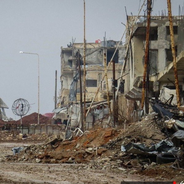 الصورة تظهر مشهدًا لمدينة مدمرة، حيث تبدو المباني متضررة بشدة، مع بقايا الهياكل المعدنية وأجزاء من الأنقاض. الطريق في المنتصف مليء بالحصى والأحجار، ويبدو غارقًا في الخراب. يظهر في الخلفية بعض الأعمدة الكهربائية التي لم تعد تعمل، مما يضيف إلى الجو القاتم والموحش للمكان. الأجواء عامة غائمة، مما يزيد من شعور الخواء واليدانة في هذا المشهد.