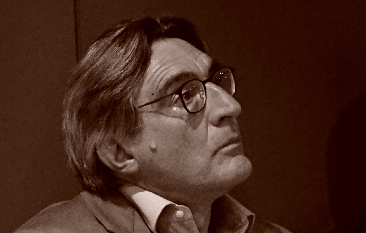 در این تصویر، فردی با موهای خاکستری و چهرهای جدی به نظر میرسد. او عینک زده و لباس روشنی بهتن دارد. در پسزمینه، دیوار تاریکی دیده میشود و به نظر میرسد که فرد در حال فکر کردن یا تماشا کردن چیزی است. تصویر دارای تن رنگی گرم و طبیعی است که به حالت contemplative او افزوده است.