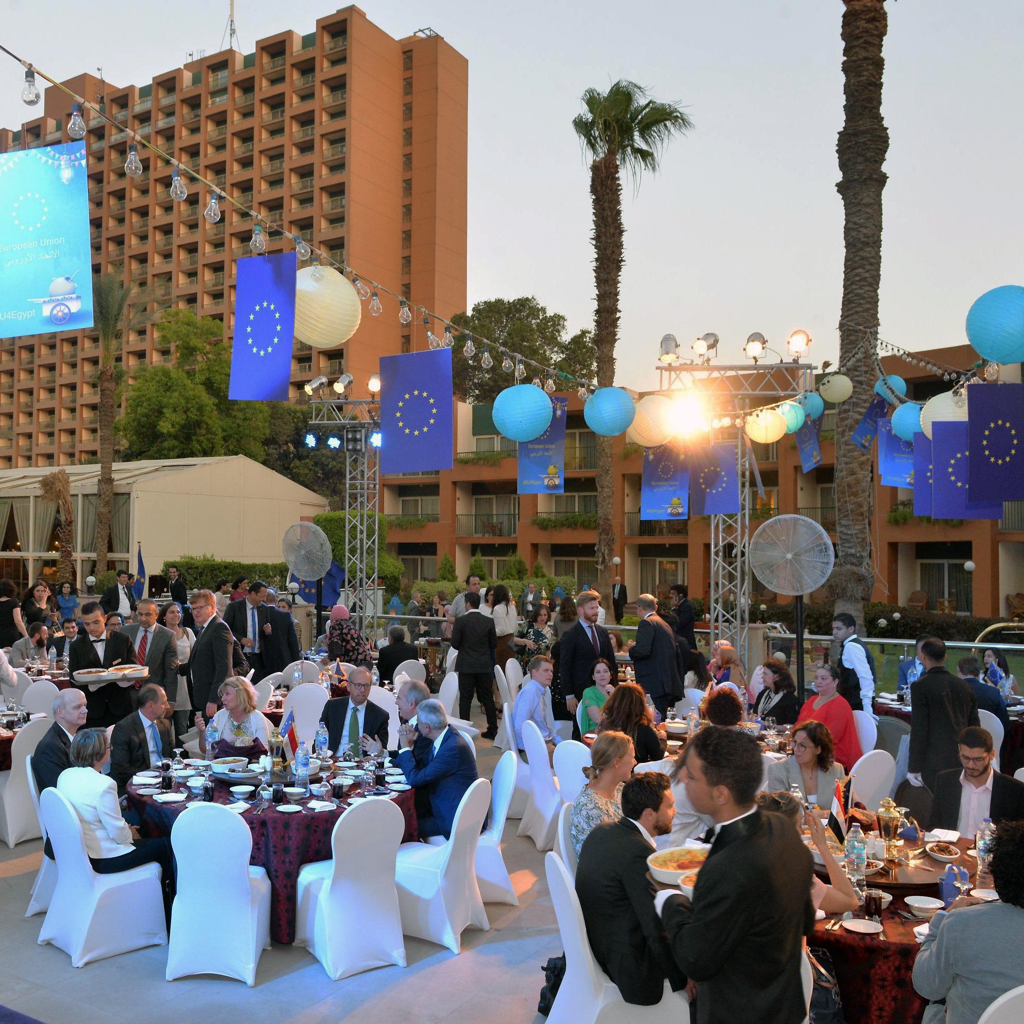 L'image montre un événement extérieur festif, probablement un dîner ou une réception. Des tables rondes sont disposées avec des nappes élégantes, où des convives en tenues formelles s'assoient pour profiter du repas. L'environnement est décoré avec des lanternes et des bannières aux couleurs de l'Union européenne. Le décor comprend également des palmiers et des installations lumineuses, créant une ambiance conviviale et festive au crépuscule. Les personnes présentes semblent converser joyeusement et profiter de l'événement.