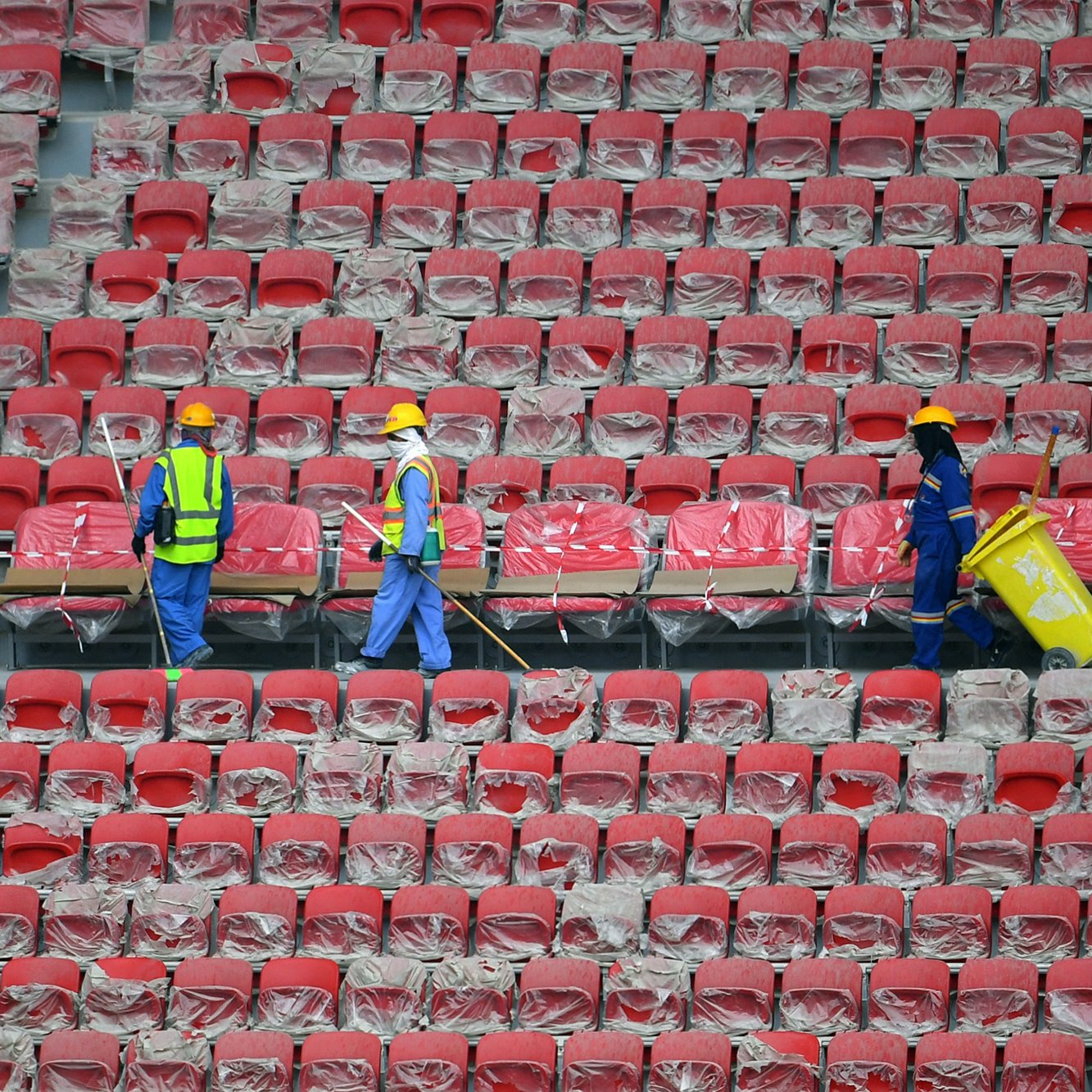 L'image montre des travailleurs dans un stade vide. Ils portent des casques de protection et des gilets réfléchissants. Les sièges du stade sont recouverts de plastique, et les travailleurs semblent être en train de nettoyer ou de préparer les lieux. On peut voir des rangées de sièges rouges. L'atmosphère paraît calme et organisée.