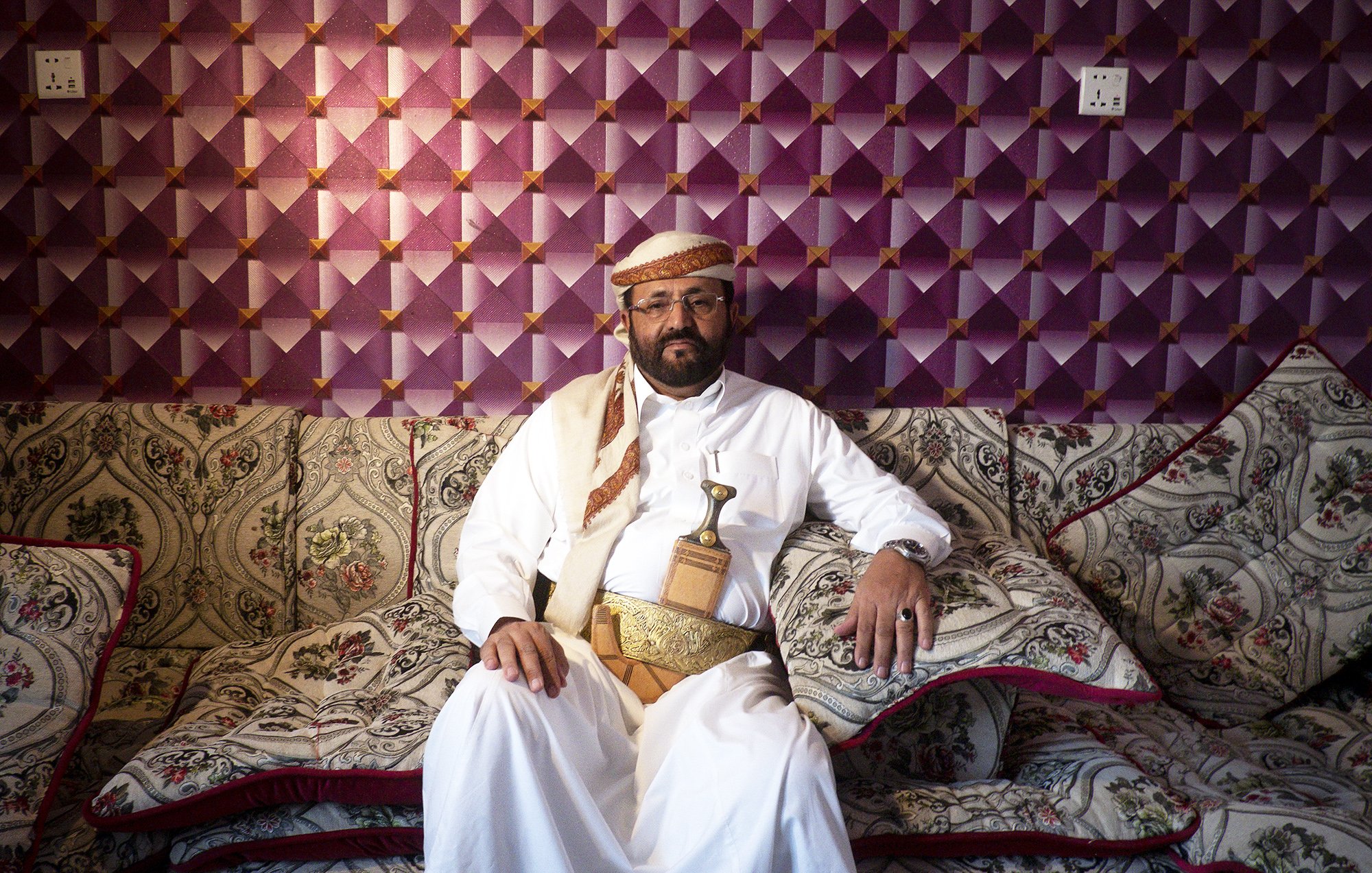 L'image montre un homme vêtu d'un habit traditionnel, assis sur un canapé orné de motifs décoratifs. Il porte une ceinture dans laquelle est insérée une sorte de poignard, typique de certaines cultures arabes. Le fond est constitué d'un mur avec un revêtement texturé en violet, ajoutant une touche colorée à la scène. L'homme a une expression calme et posée, et son environnement reflète une atmosphère accueillante et traditionnelle.