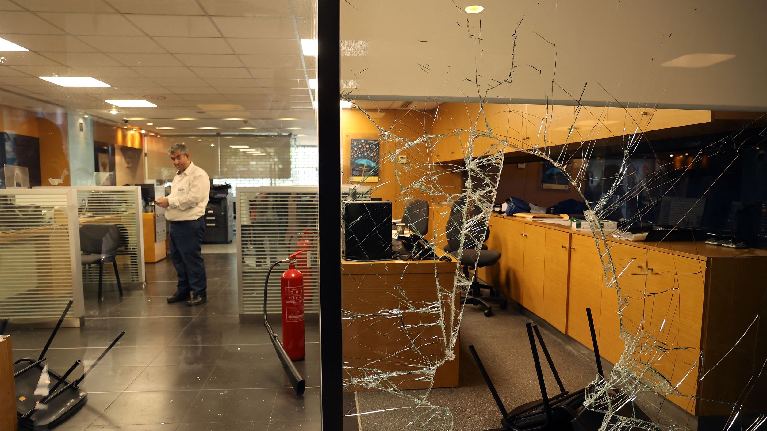 این تصویر یک نمای داخلی از یک دفترکار را نشان می‌دهد که به وضوح آسیب دیده است. شیشه‌ی پنجره شکسته و تکه‌های آن روی زمین پخش شده‌اند. در داخل اتاق چند صندلی معکوس و به هم ریخته وجود دارد. یک مرد در حال ایستادن در نزدیکی کامپیوترها به نظر می‌رسد و مشغول بررسی وضعیت است. همچنین در گوشه‌ای از اتاق، یک کپسول آتش‌نشانی قرار دارد. فضای کلی تصویر حاکی از بی‌نظمی و آسیب‌های شدید در این محیط کاری است.