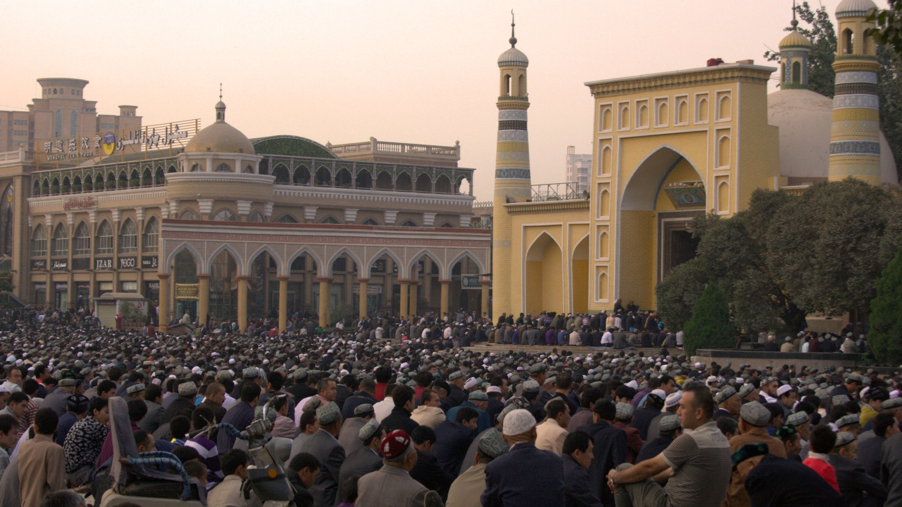 تش depicts الصورة تجمعًا كبيرًا من الناس، حيث يجلس عدد كبير من الأفراد في ساحة مفتوحة. في الخلفية، يمكن رؤية معمار تقليدي يتضمن مآذن وقباب، مما يشير إلى أنه قد يكون موقعًا دينيًا. السماء في حالتها النهارية تكتسي بألوان دافئة، مما يضفي جواً خاصاً على المشهد. يبدو أن الناس مجتمعون لأداء نشاط جماعي، ربما صلاة أو احتفال.