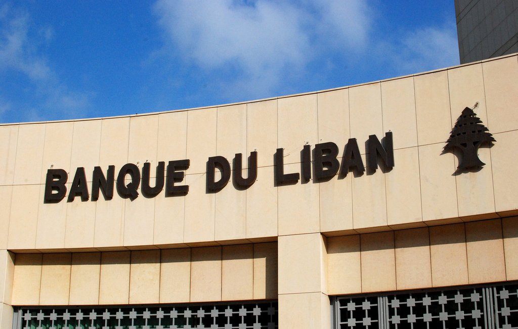 L'image montre un bâtiment avec l'inscription "BANQUE DU LIBAN" en lettres majuscules. La façade est faite de pierre ou de béton, et il y a un symbole d'un petit arbre à côté du texte. Le ciel en arrière-plan est bleu avec quelques nuages. L'ensemble donne une impression de modernité et de solidité.