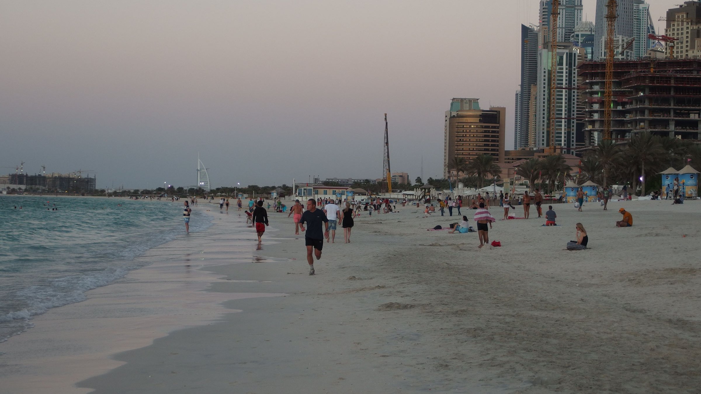 L'image montre une plage avec des personnes marchant le long du rivage. On aperçoit des baigneurs et des groupes assis sur le sable. En arrière-plan, on distingue des bâtiments modernes et des gratte-ciel, symboles d'une grande ville, ainsi que des grues témoignant de travaux de construction. Le ciel présente des teintes douces, probablement au moment du crépuscule, créant une atmosphère paisible et attractive. Les vagues de la mer semblent calmes, ajoutant à la sérénité du paysage.