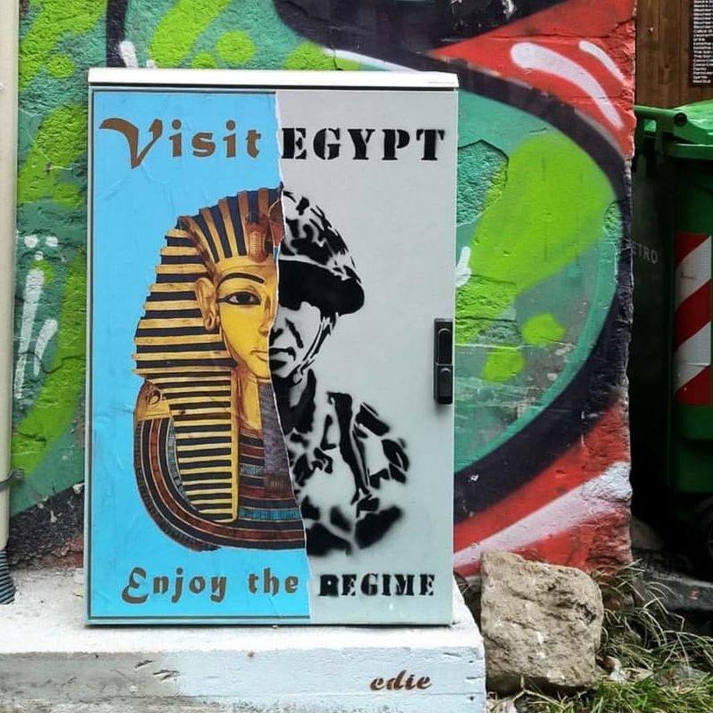 L'immagine mostra un armadietto metallico dipinto con un murale che presenta due volti distinti. A sinistra, c'è un'immagine stilizzata di un faraone, accompagnata da un invito a "Visit Egypt" e "Enjoy the". A destra, c'è un ritratto che sembra rappresentare un soldato o una figura autoritaria, con la parola "REGIME" sopra di esso. Lo sfondo è colorato e impreziosito da graffiti, creando un contrasto tra i due soggetti. La composizione sembra riflettere una critica sociale o politica legata all'Egitto.
