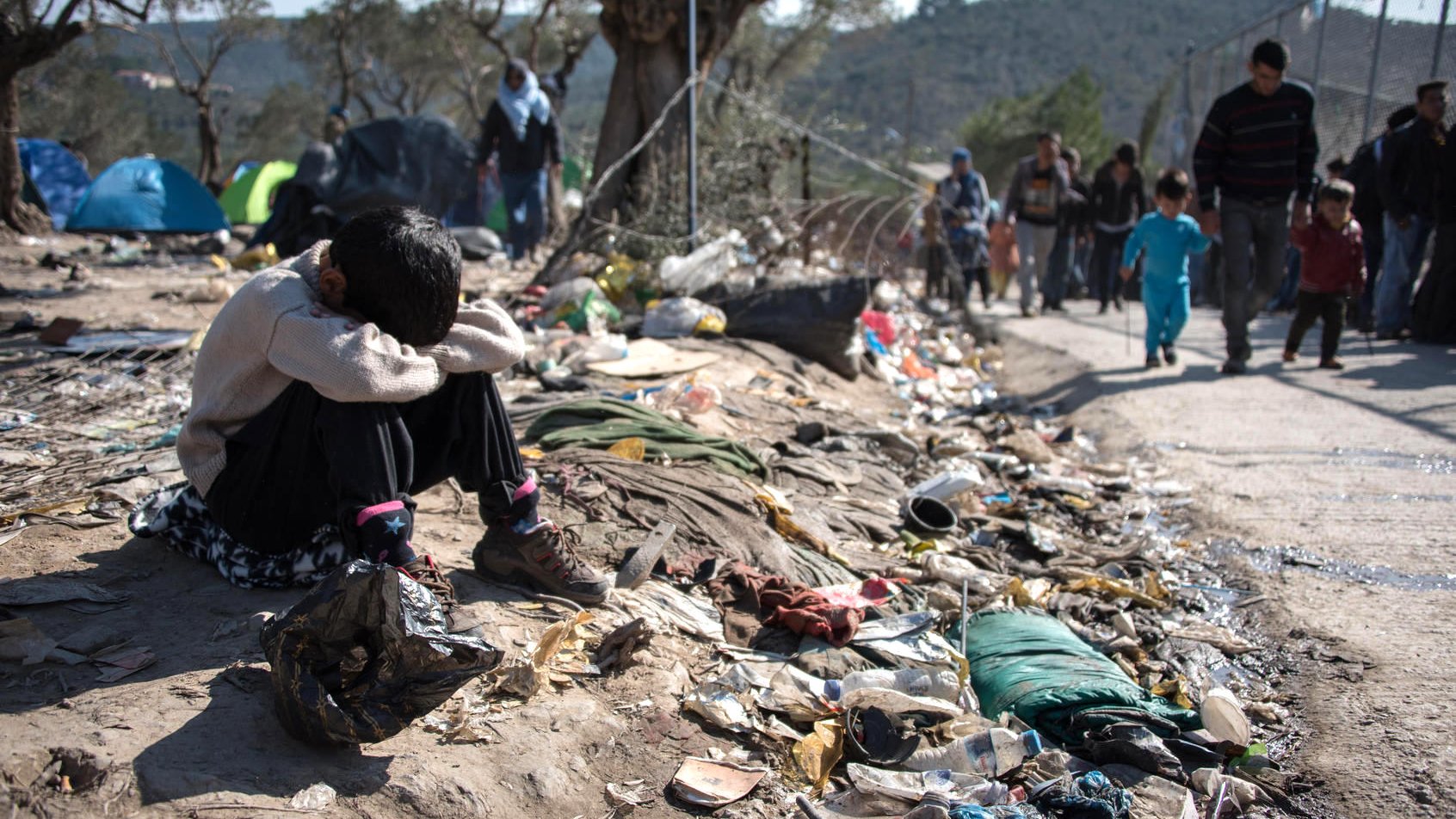 تظهر الصورة طفلاً يجلس على الأرض في ملجأ مهدم أو منطقة غير نظيفة، حيث يبدو عليه الإحباط أو الحزن، ويضع رأسه بين ركبتيه. حوله هناك أكوام من القمامة والأشياء المهملة، مما يعكس ظروف معيشة قاسية. في الخلفية، يمكن رؤية مجموعة من الأشخاص تسير في اتجاه آخر، مما يعزز الشعور بالانفصال والمعاناة. تعكس الصورة بوضوح التحديات التي يواجهها الأفراد في مثل هذه الظروف.