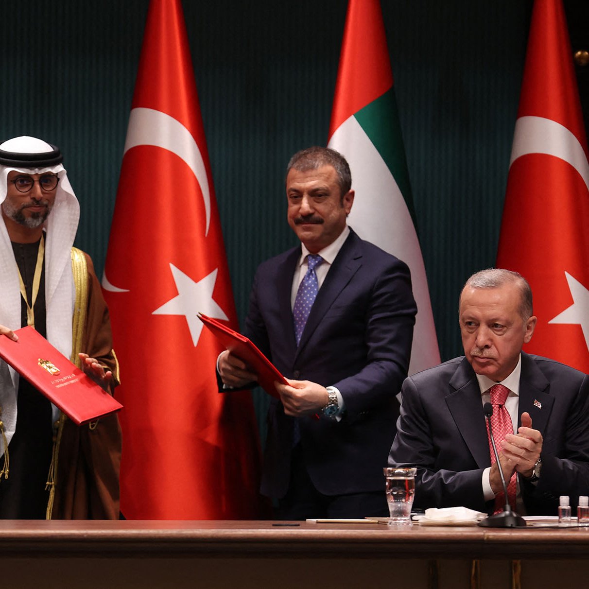 L'immagine mostra una cerimonia ufficiale con quattro uomini in abiti formali. Sullo sfondo si vedono le bandiere degli Emirati Arabi Uniti e della Turchia. Due degli uomini sono in piedi e sembrano scambiarsi documenti, mentre gli altri due sono seduti. L'atmosfera suggerisce un momento importante, probabilmente relativo a un accordo o a una collaborazione tra i due paesi.