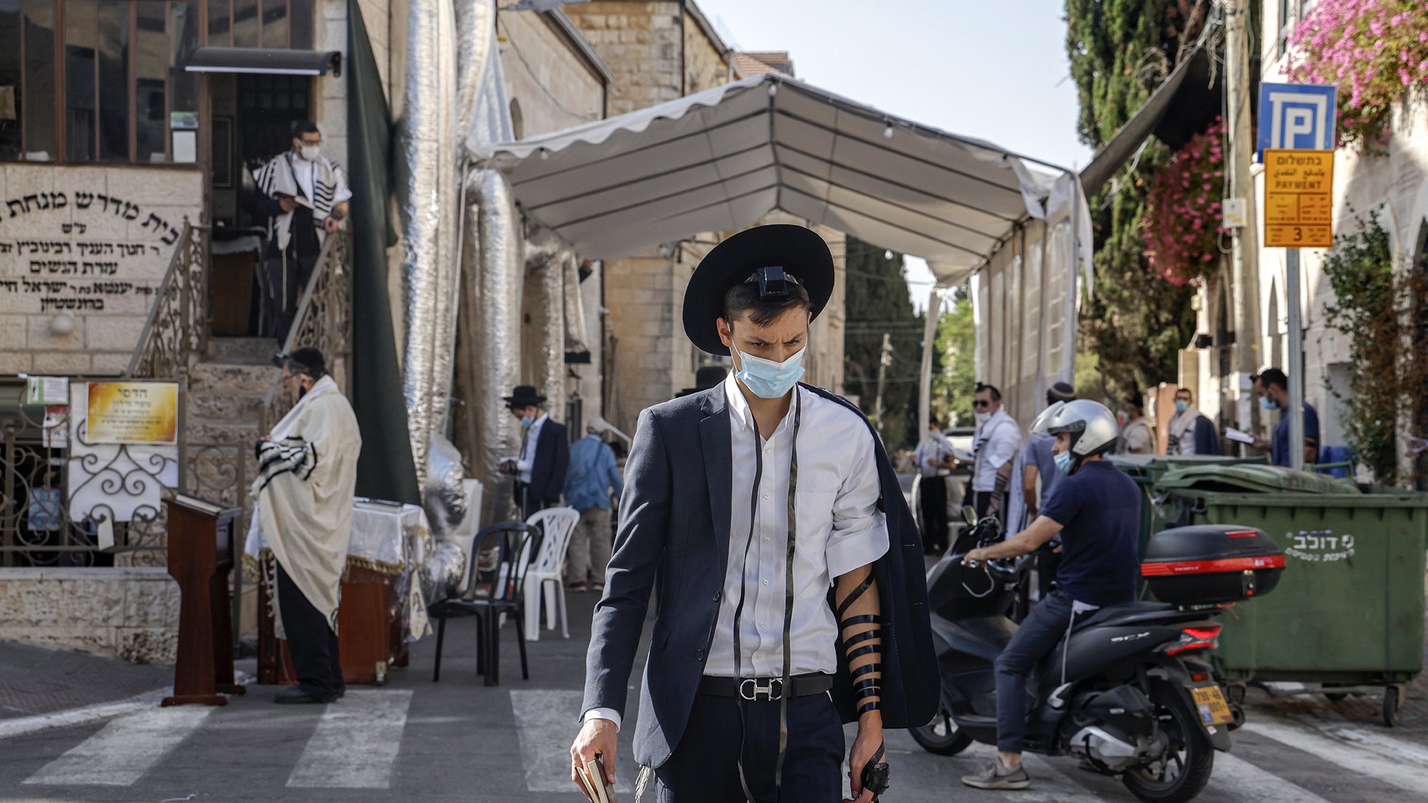 En la imagen se puede ver a un hombre joven que lleva puesta una camisa blanca y un saco oscuro, además de un sombrero negro. Está caminando por una calle que parece estar cerca de un lugar de culto judío, ya que al fondo se observa un edificio con elementos relacionados con la cultura y religión judía. El hombre lleva una máscara, posiblemente debido a las precauciones sanitarias. Se puede notar que tiene filacterias (tefilín) en su brazo, lo cual es parte de la práctica judía durante las oraciones. A su alrededor hay otras personas, algunas en la acera y otras en vehículos, lo que sugiere que es un lugar activo. Hay también un toldo montado, probablemente utilizado para eventos religiosos.