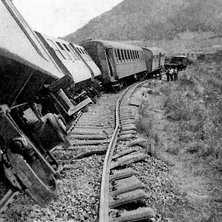 L'image montre un accident de train. Plusieurs wagons sont déraillés, penchés sur le côté, tandis que les rails semblent déformés. En arrière-plan, on aperçoit des montagnes et des personnes qui semblent inspecter les lieux. L'atmosphère est celle d'un événement tragique, évoquant une situation d'urgence.