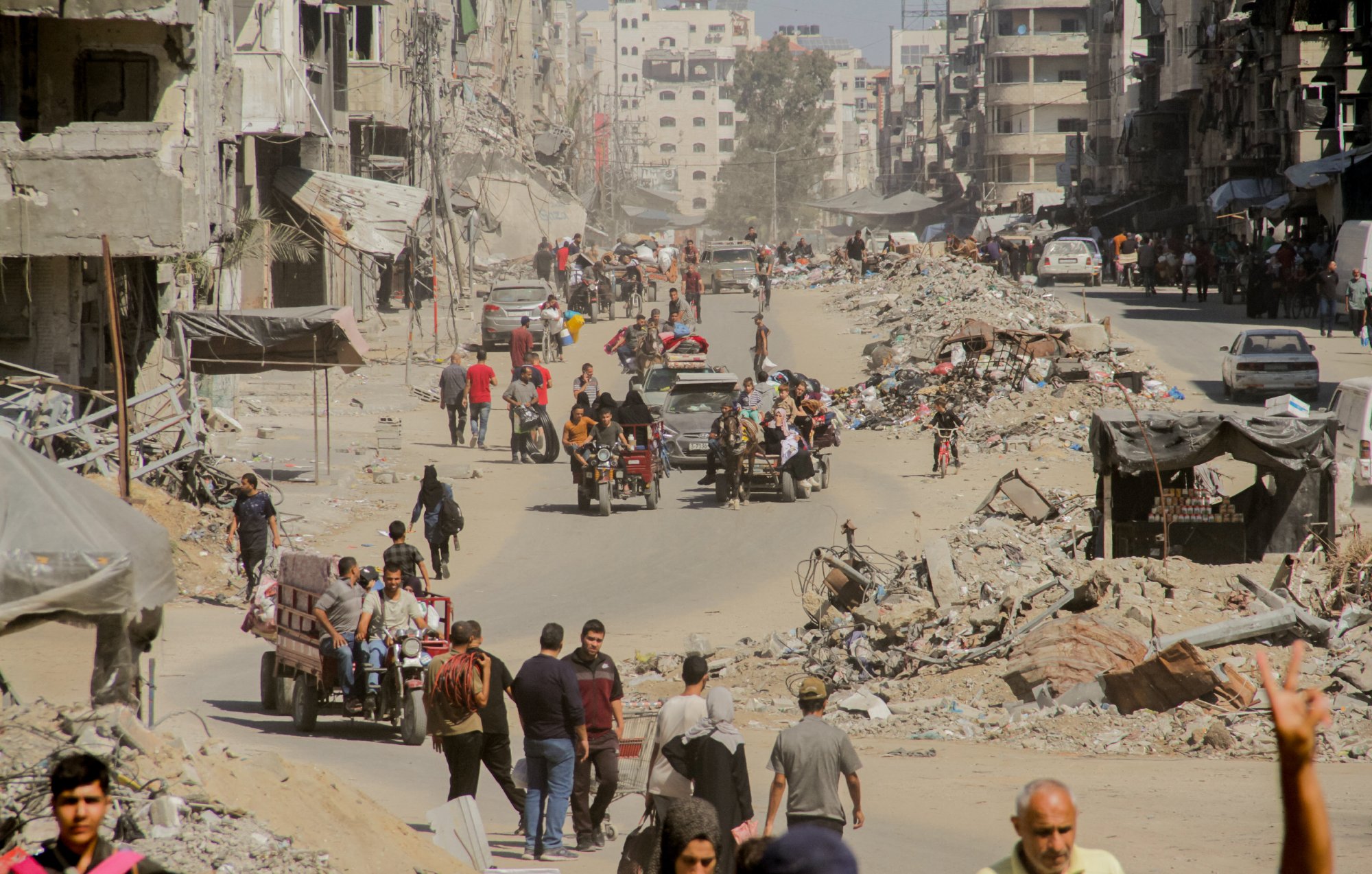 L'immagine mostra una scena di una città con edifici distrutti e rovine visibili. La strada è affollata da persone che camminano e da veicoli, inclusi motocicli e carretti. La polvere sollevata dall'ambiente circostante crea un'atmosfera di desolazione. In lontananza si vedono ulteriori segni di distruzione, con detriti sparsi lungo i lati della strada. L'insieme comunica un forte senso di difficoltà e resilienza in un contesto di crisi.
