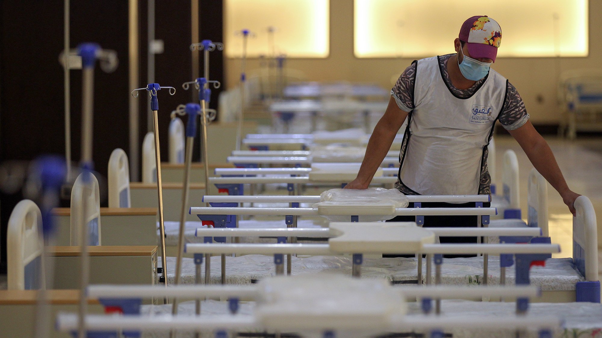 در این تصویر، یک مرد در حال کار در یک فضای بیمارستانی یا درمانگاهی دیده می‌شود. او یک ماسک به صورت دارد و به تخت‌هایی که در ردیف‌های مرتب قرار دارند، توجه می‌کند. تخت‌ها با ملحفه‌های سفید پوشیده شده‌اند و تعدادی از آن‌ها به دستگاه‌های اکسیژن متصل هستند. نور محیط ملایم است و فضا به نظر تمیز و سازمان‌دهی شده می‌رسد.