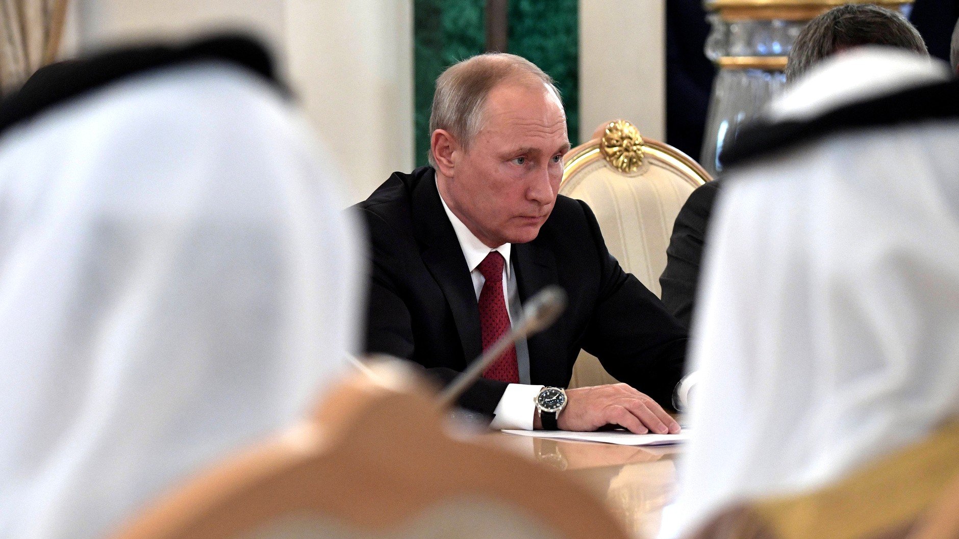 تظهر الصورة اجتماعًا رسميًا حيث يجلس رجل يرتدي بدلة ذات لون داكن. يبدو أنه مركز جداً، حيث ينظر إلى الطاولة أمامه. في الخلفية، يمكن رؤية أشخاص آخرين يرتدون الملابس التقليدية، مما يعطي انطباعًا عن حدث رسمي أو دبلوماسي يشارك فيه ممثلون من ثقافات مختلفة. يحيط بالاجتماع أجواء من الجدية والتركيز.