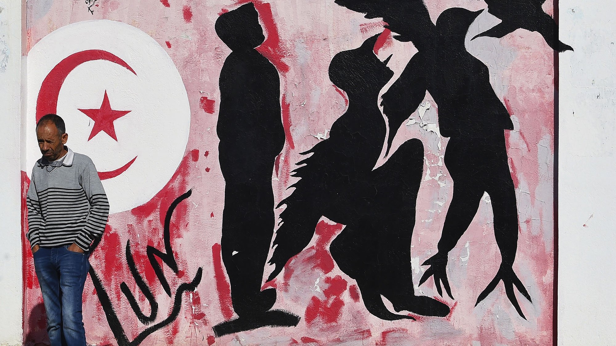 L'image montre un mur peint avec des graffitis. À gauche, on peut voir le drapeau tunisien en rouge et blanc, avec un croissant et une étoile. À côté, des silhouettes humaines sont représentées en noir sur un fond rose, certaines semblent interagir avec des éléments d'oiseaux, suggérant des thèmes de liberté ou de mouvement. Un homme en pull rayé marche le long du mur, ajoutant un aspect vivant à la scène.