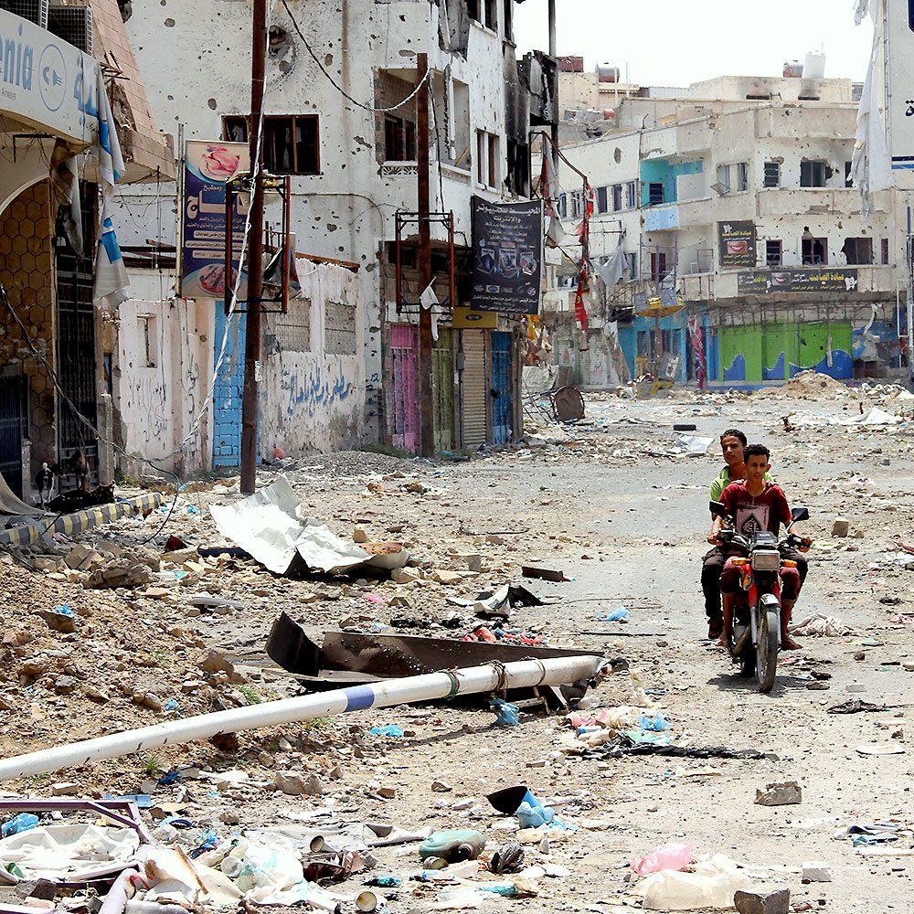 تظهر الصورة مشهداً لشارع في منطقة مهجورة أو متضررة، حيث تنتشر الفوضى والخراب. الشارع مليء بالقمامة والحطام، والمباني المحيطة تبدو متدهورة ومهجورة، مع بعض العلامات الإرشادية المتبقية. في وسط الصورة، يوجد شخصان يجلسان على دراجة نارية، مما يضيف لمسة من الحياة إلى المشهد القاتم. يبدو أن المكان يفتقر إلى الحركة والحيوية، مما يعكس حالة من الإهمال.