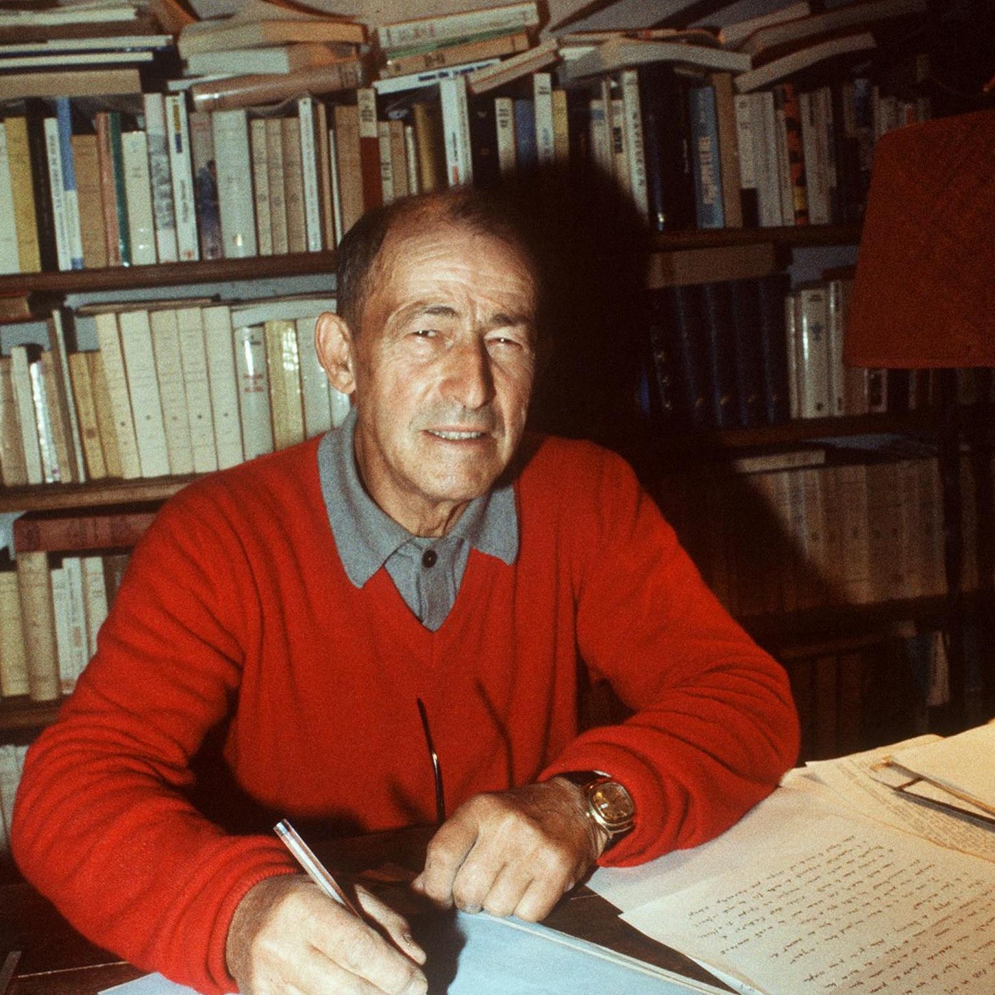 L'image montre un homme assis à un bureau, souriant, en train d'écrire sur une feuille de papier avec un stylo. Il porte un pull rouge et une chemise à col. En arrière-plan, on peut voir une bibliothèque remplie de livres. Sur le bureau, il y a également des feuilles de papier, peut-être des notes ou un manuscrit en cours de rédaction. L'éclairage crée une ambiance chaleureuse et studieuse.