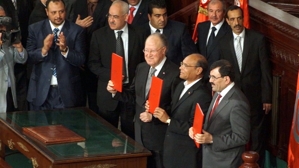 L'image montre un groupe d'hommes en costume, apparemment dans un cadre officiel, probablement lors d'une cérémonie ou d'un événement politique. Ils tiennent des documents en main, et l'ambiance semble solennelle. On peut voir une table en bois devant eux ainsi que des drapeaux en arrière-plan. Les personnes présentes semblent applaudir ou se concentrer sur l'événement.