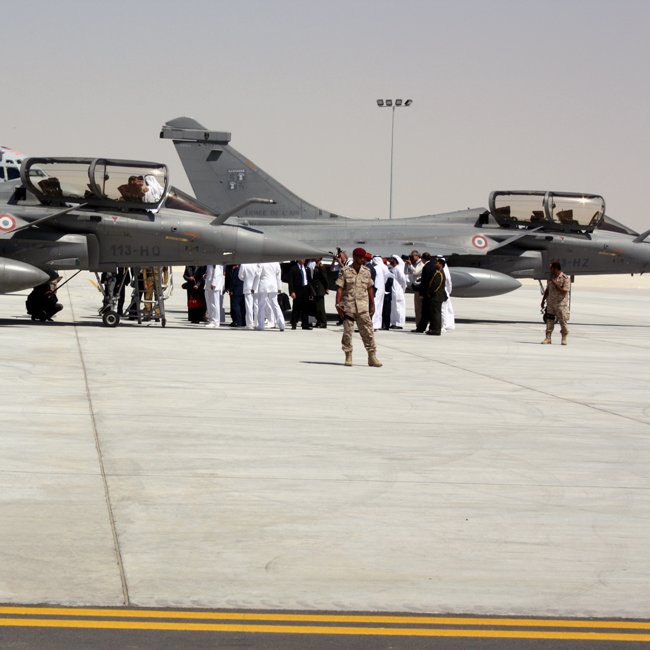 L'image montre une scène sur un tarmac d'aéroport militaire. On y voit plusieurs avions de chasse, probablement des Mirage ou des jets similaires, garés sur une surface bétonnée. Des membres d'équipage, habillés en uniforme, interagissent avec des personnes portant des costumes traditionnels. En arrière-plan, il y a d'autres personnes rassemblées, ce qui suggère un événement ou une cérémonie. Le cadre est désert, avec un ciel clair, ce qui indique qu'il pourrait s'agir d'une région aride.