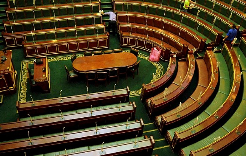 تظهر الصورة قاعة اجتماعات برلمانية، حيث تتوزع المقاعد بشكل دائري حول طاولة مستطيلة في المنتصف. المقاعد مصممة بطريقة تتسع لعدد كبير من الأعضاء، بينما يبدو الجو هادئًا. بعض العمال يقومون بتنظيف القاعة، مما يوحي بأن الاجتماع قد انتهى أو لم يبدأ بعد. الأرضية مزينة بنمط معين، مما يضيف جمالية للمكان.