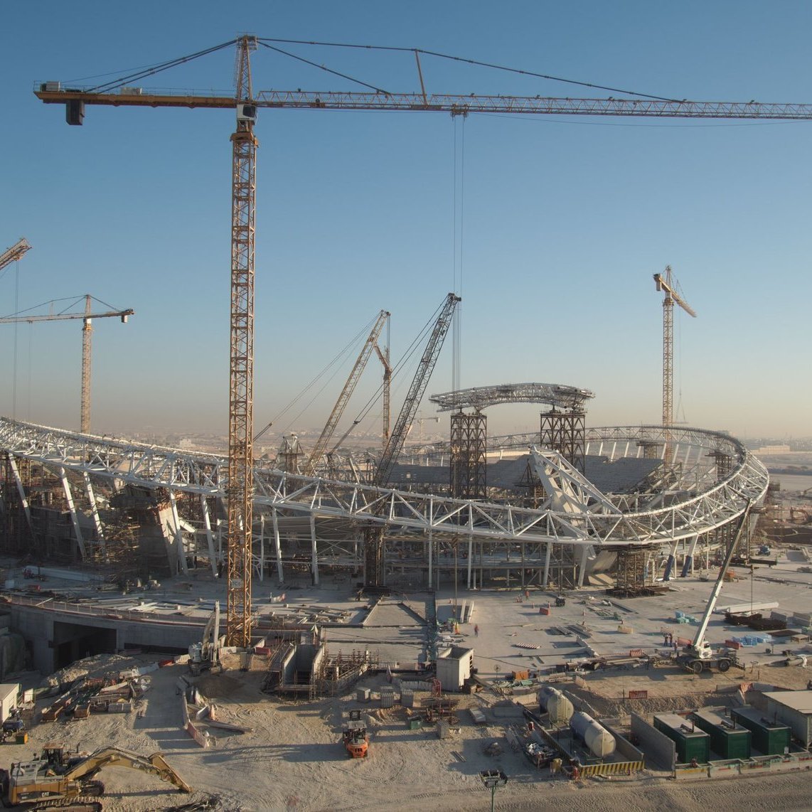 L'image montre un grand chantier de construction d'un stade, avec plusieurs grues en fonctionnement. Le stade a une structure moderne et architecturale, avec des éléments en acier visibles. En arrière-plan, on peut voir un ciel clair, suggérant qu'il fait jour. Le site est entouré de divers matériaux de construction et d'équipements, ce qui indique que le projet est encore en cours.