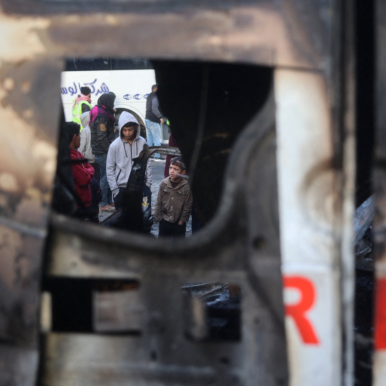 تصویر نشان‌دهنده‌ی یک صحنه‌ی غم‌انگیز است که در آن چندین کودک و بزرگسال درحال مشاهده‌ی یک وضعیت بحرانی به نظر می‌رسند. در پس‌زمینه، یک وسیله نقلیه آسیب‌دیده دیده می‌شود که حالت سوخته و خراب دارد. نام "پرس" بر روی آن به وضوح مشاهده می‌شود. در داخل نمای این وسیله، افرادی با حالت‌هایی جدی و نگران دیده می‌شوند که شاید به دنبال کمک یا راهی برای فرار از وضعیت خطرناک باشند. احساس تنش و اضطراب در این تصویر محسوس است.
