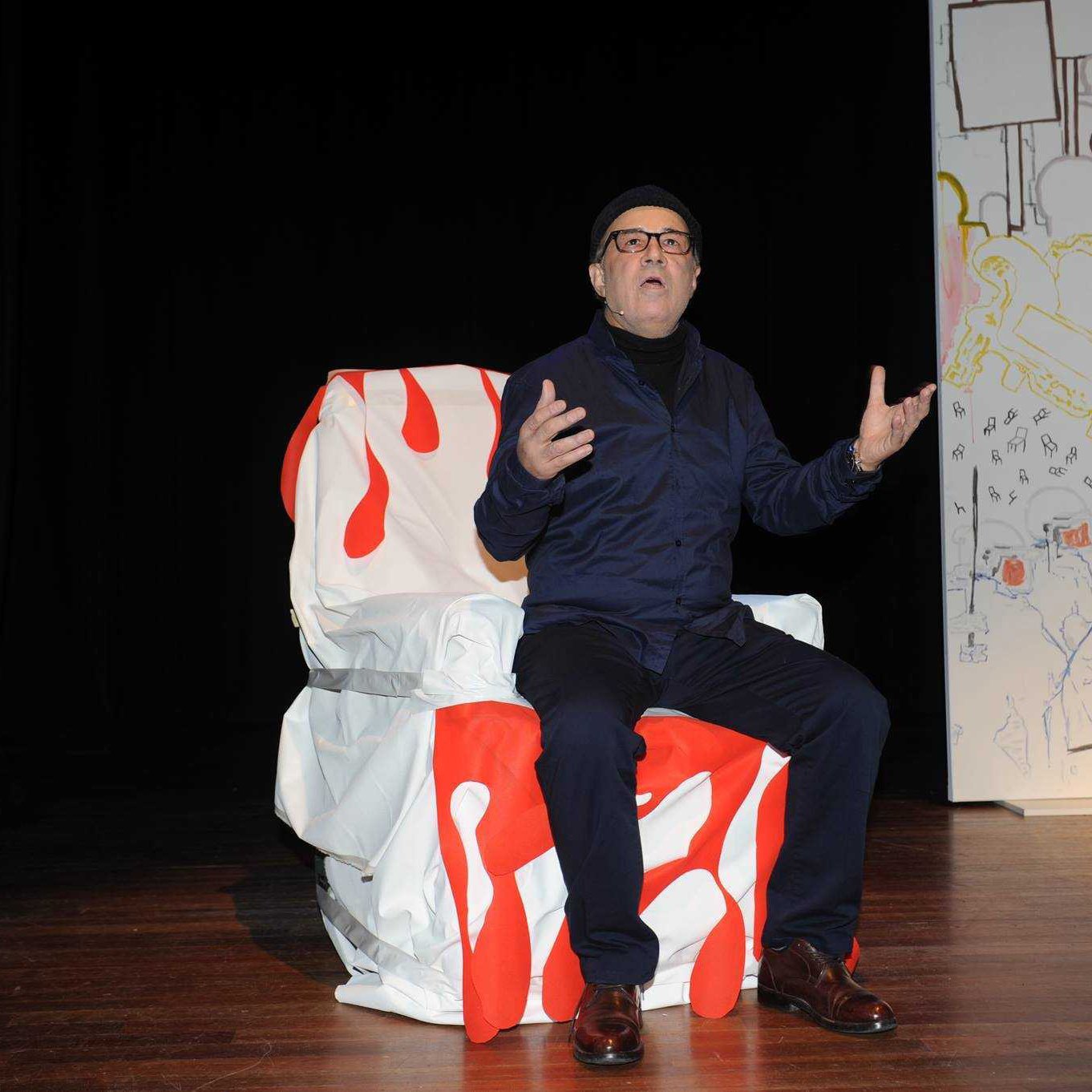 تظهر في الصورة شخصية جالسة على كرسي مغطى بقماش أبيض مزين بلمسات حمراء. يبدو أنه في وضع أداء أو حديث، حيث يعبّر باستخدام يديه ويظهر تركيزاً في تعبيراته. الخلفية تحتوي على لوحات جدارية غير محددة، مما يضفي جواً فنياً على المشهد. الشخص يرتدي ملابس داكنة ونظارات، ويبدو بشكل عام في حالة تفاعل مع الجمهور أو المشهد المحيط به.