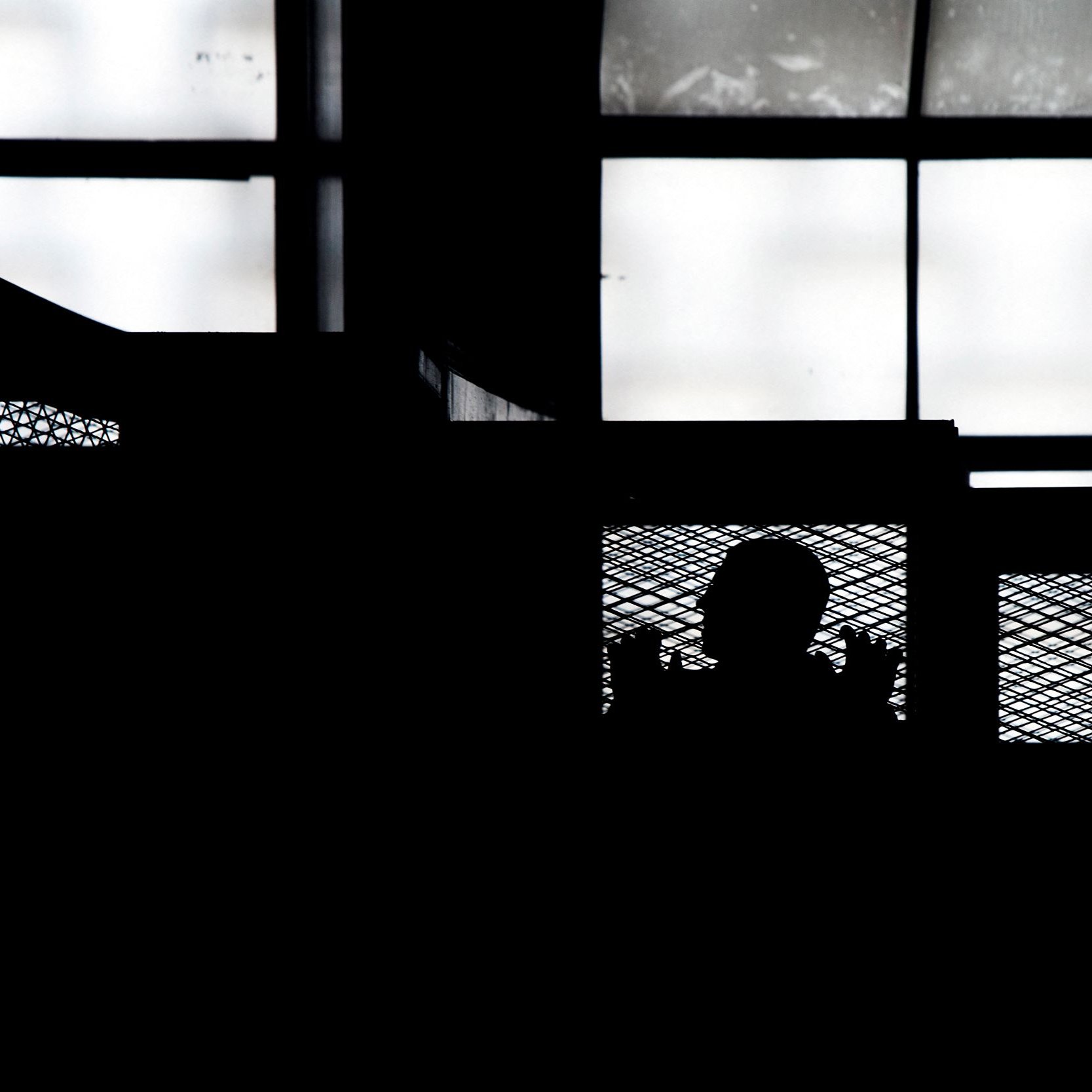 L'image montre une silhouette, probablement d'une personne, derrière une structure en métal qui semble être une grille ou une barrière. En arrière-plan, on aperçoit des fenêtres laissant passer une lumière diffuse. L'ensemble de la scène est dominé par des ombres, créant une atmosphère mystérieuse. Les contours des objets sont flous, ce qui accentue l'aspect dramatique de l'image.