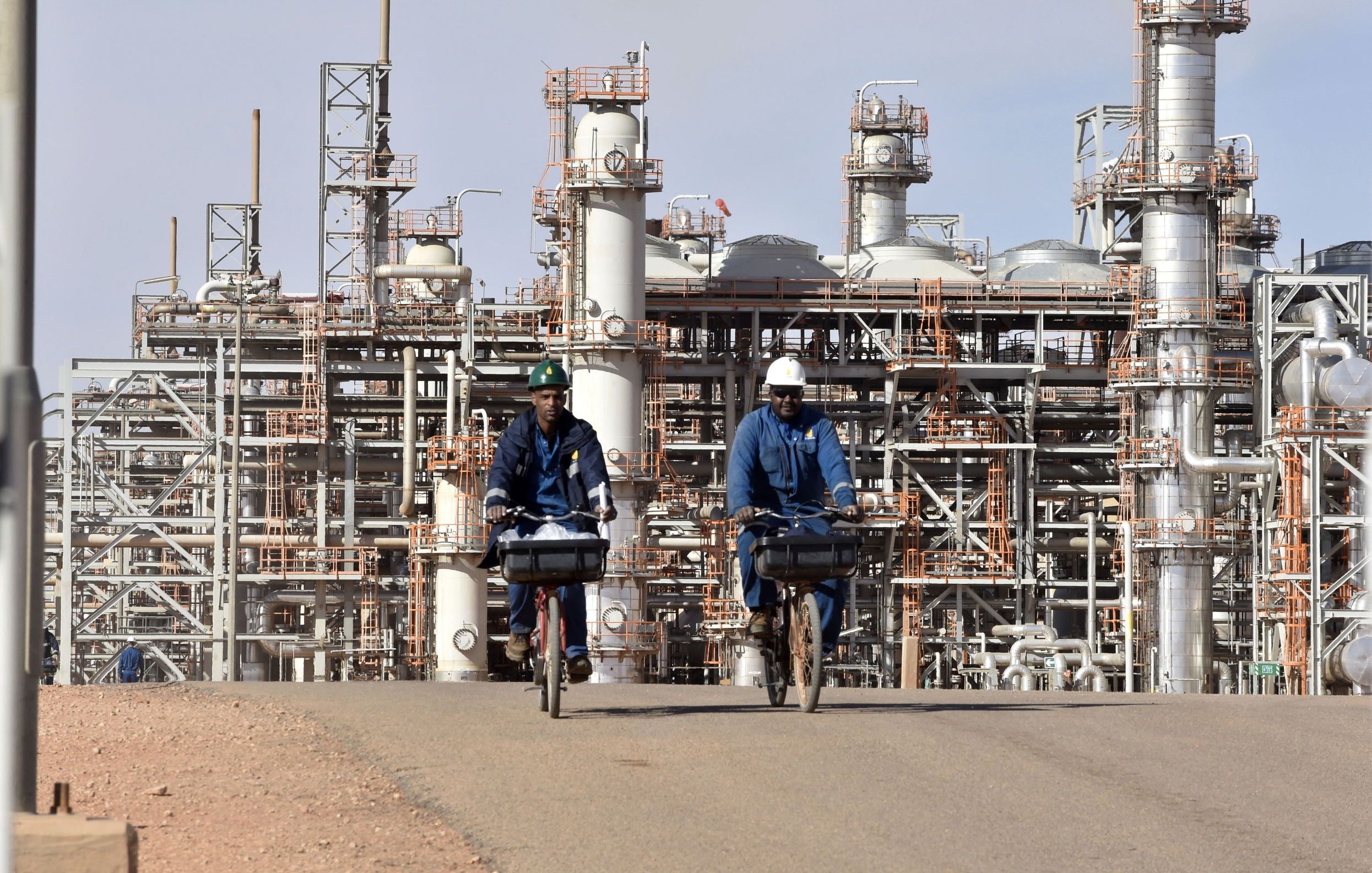 تظهر الصورة مشهدًا من منطقة صناعية، حيث يقوم شخصان يرتديان زي العمل و الخوذة بركوب دراجتين على طريق داخل المصنع. في خلفية الصورة، توجد معدات صناعية كبيرة أنابيب وأبراج مخصصة لعمليات التصنيع أو التكرير. السماء تبدو صافية والجو العام يوحي بنشاطات صناعية قوية.